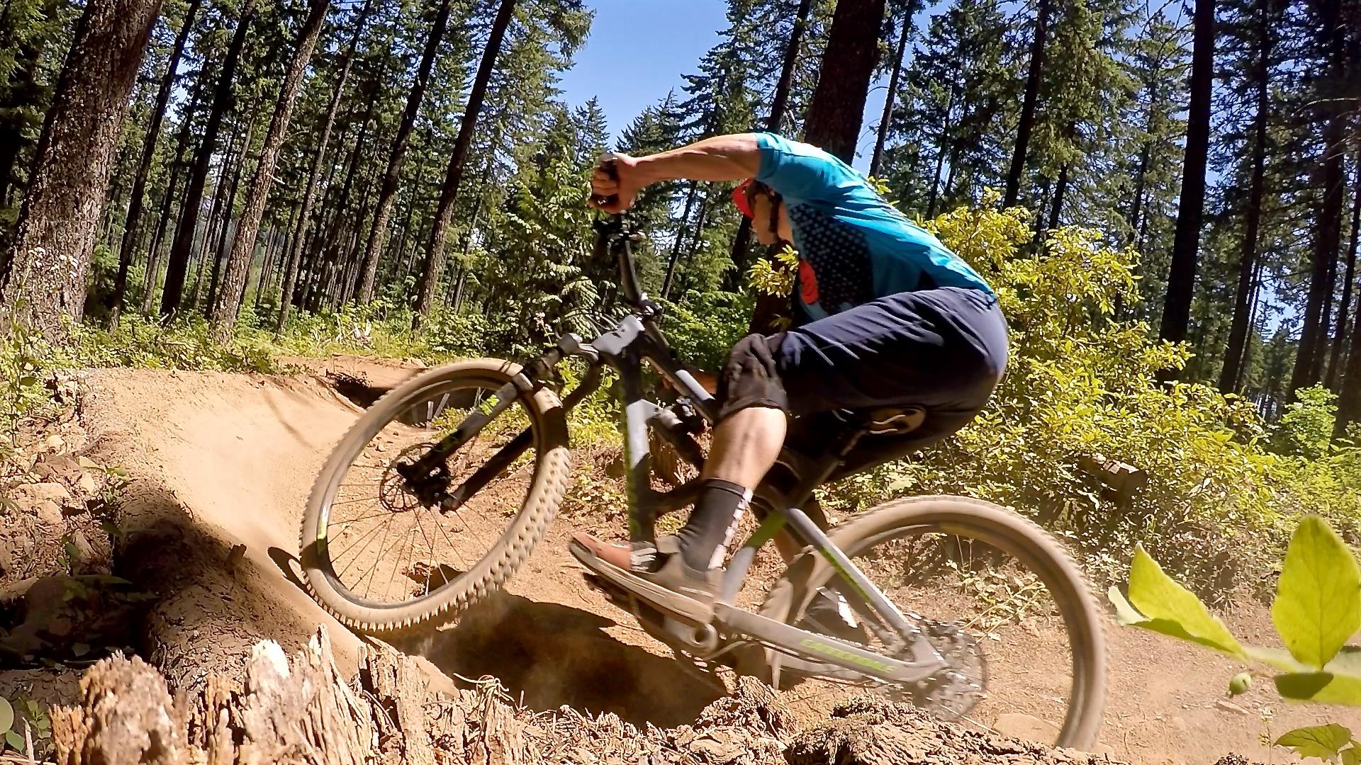 A mountain biker navigating a dirt trail through a forest, leaning into a sharp turn and kicking up dust. The rider is wearing a blue shirt and black shorts, with tall trees and green foliage in the background. Post Canyon mountain bike trail.