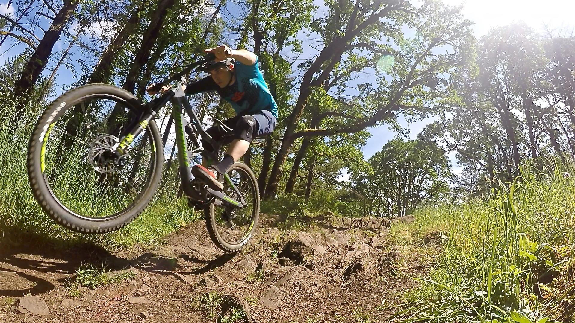 Cannondale Trigger: A mountain biker performing a jump on a rocky trail surrounded by trees and greenery, with sunlight filtering through the foliage. The cyclist is wearing a blue shirt and protective gear, showcasing a dynamic action pose as the bike’s front wheel is lifted off the ground.
