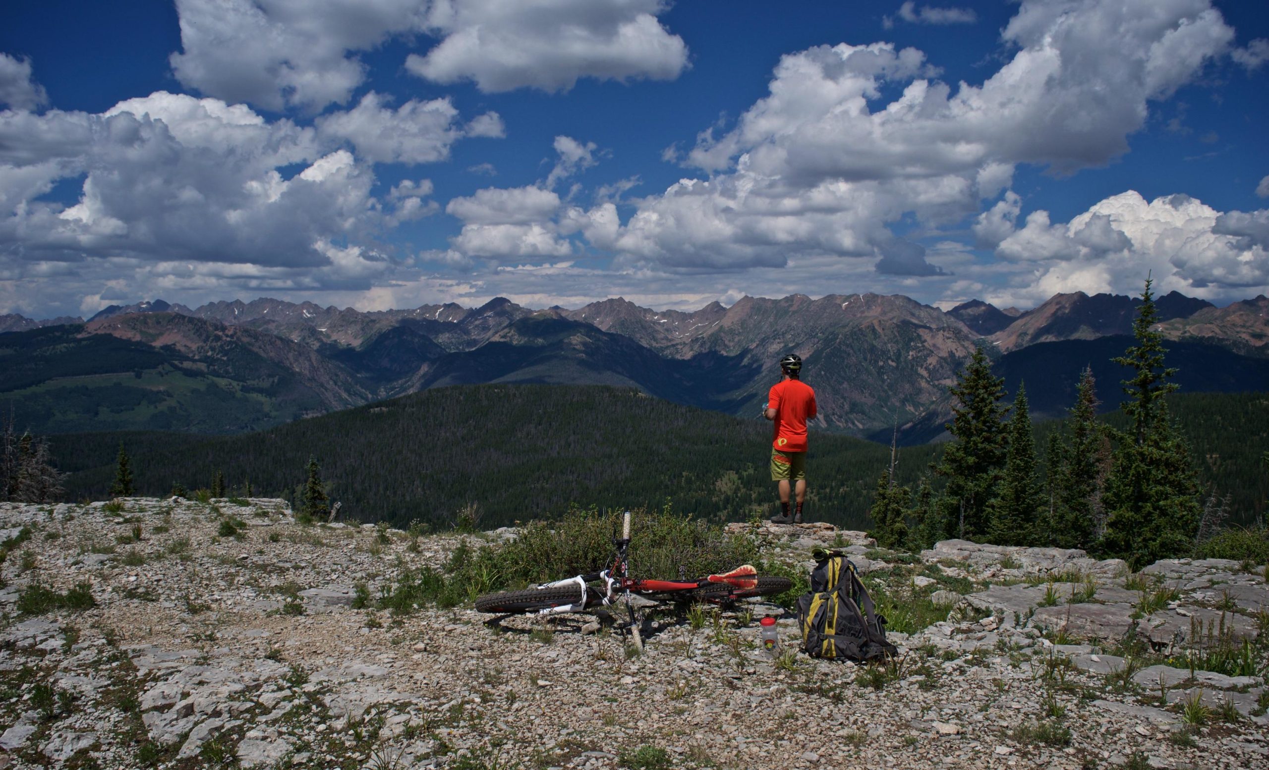 A person wearing a red shirt and shorts stands on a rocky overlook, gazing at a panoramic view of mountains and valleys under a partly cloudy sky. A mountain bike and a backpack are positioned nearby, surrounded by greenery and rocky terrain. Two Elk via Vail Pass mountain bike trail.