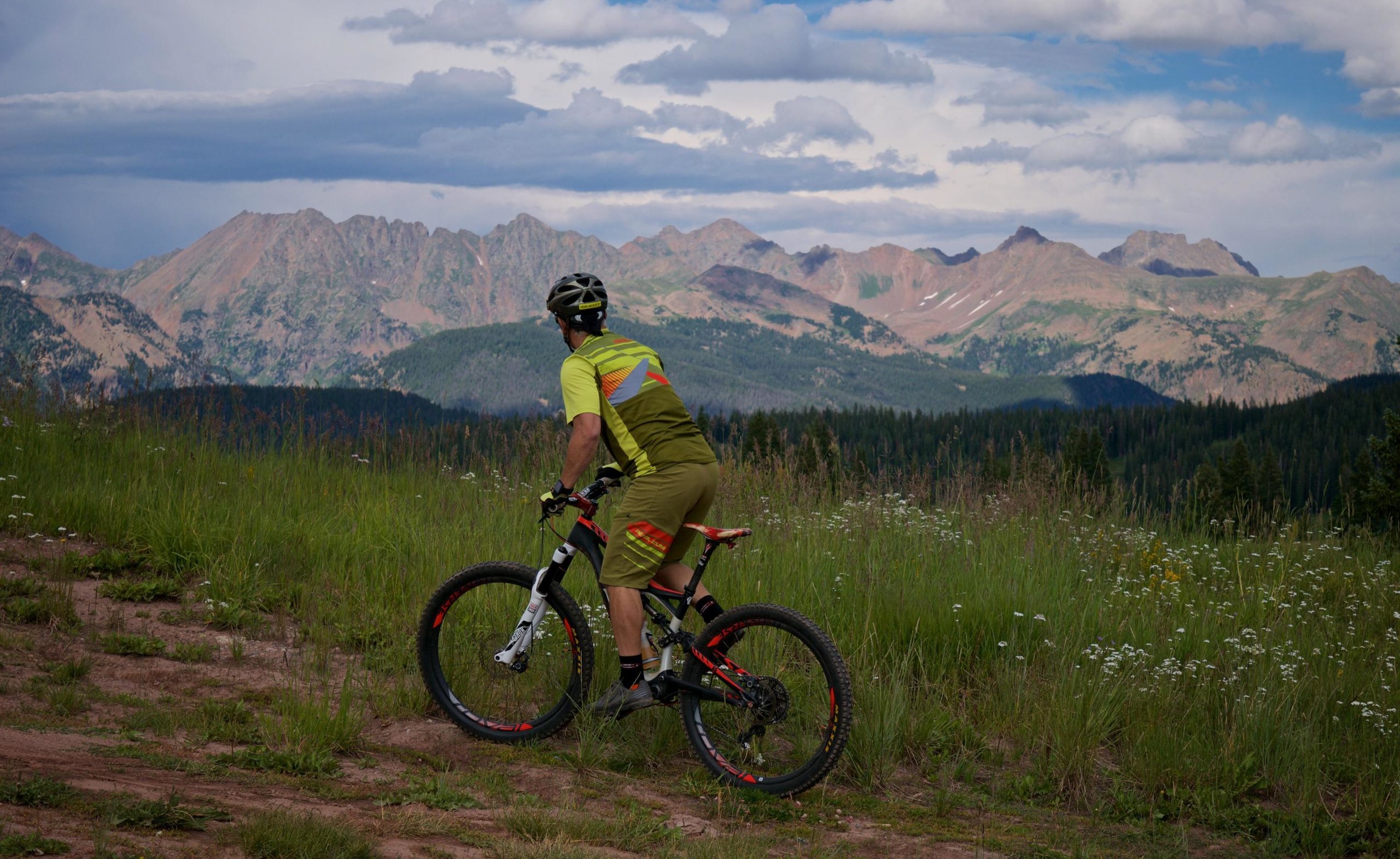 A mountain biker wearing a bright yellow and green cycling outfit is stopped on a dirt trail, gazing at the picturesque mountains in the background. Lush green grass and wildflowers surround the scene, with a partly cloudy sky above. Vail Mountain Bike Park mountain bike trail.
