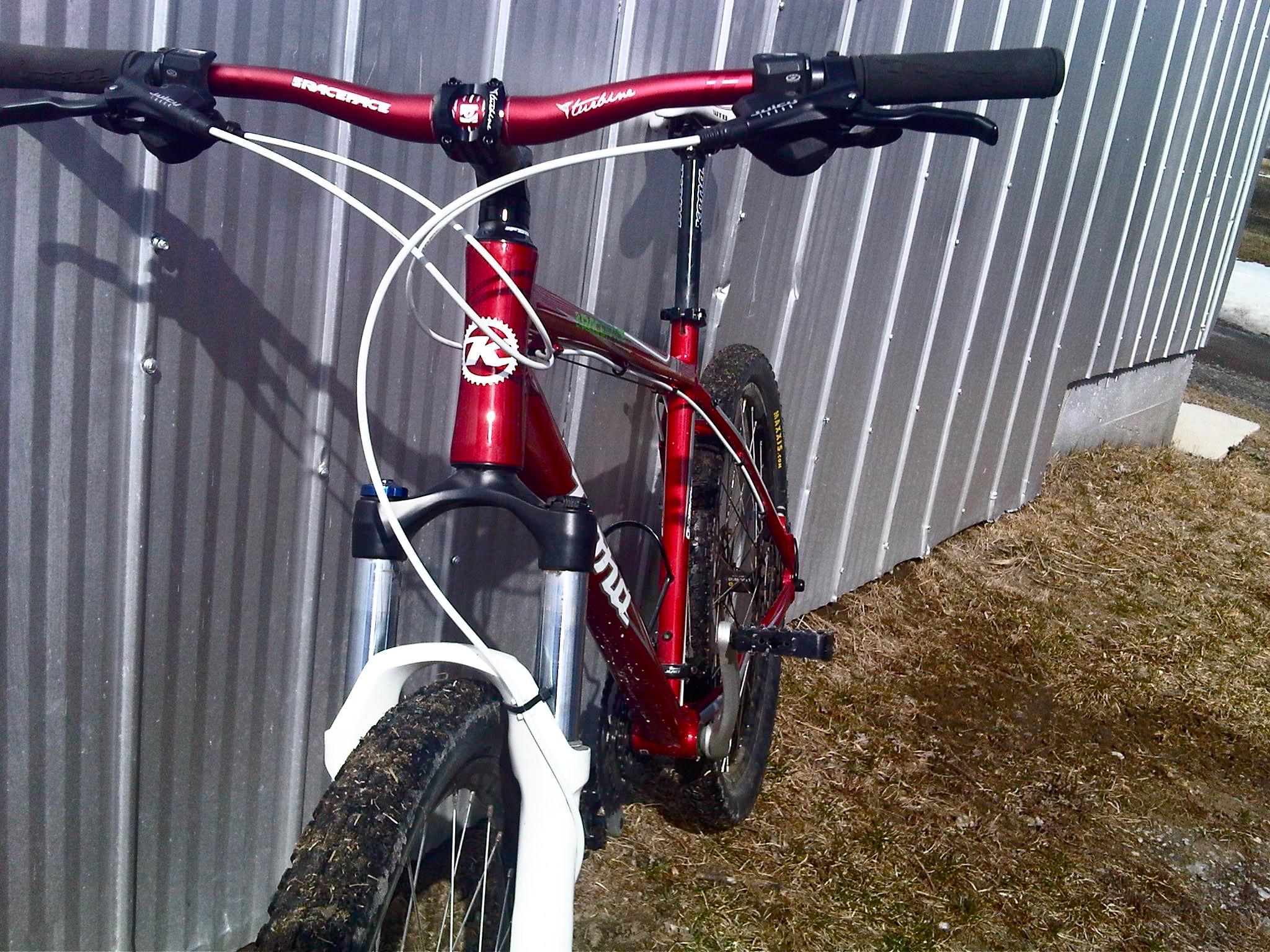 Kona Cinder Cone: A close-up view of a red mountain bike leaning against a corrugated metal wall. The bike features prominent handlebars, a front suspension fork, and thick tires with visible dirt, indicating recent use. The surrounding area includes patches of grass and a paved surface in the background.
