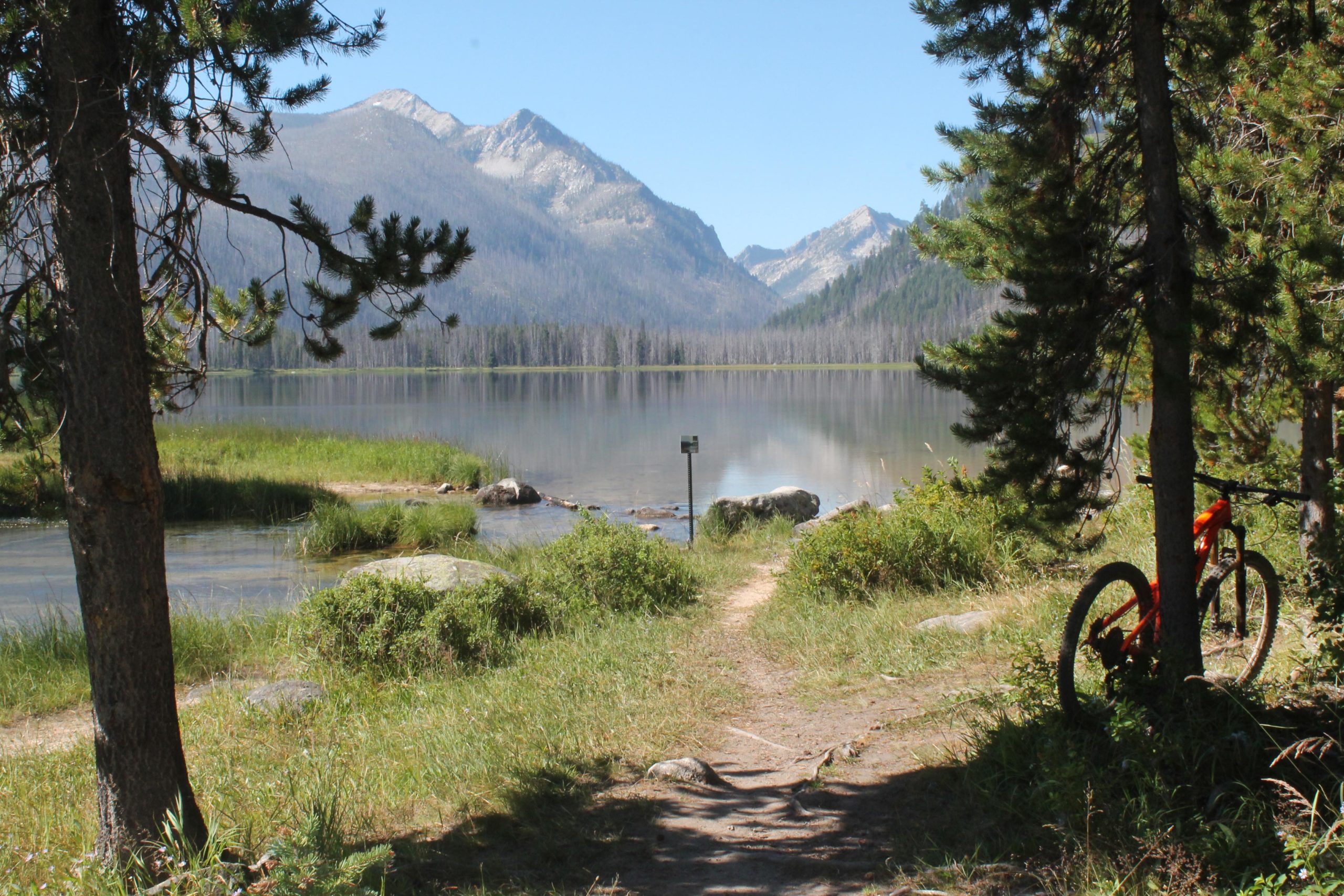 A serene landscape featuring a calm lake surrounded by mountains, with a dirt path leading to the water's edge. Tall pine trees frame the scene, and an orange mountain bike is leaning against a tree on the right. The clear blue sky reflects on the lake’s surface, creating a peaceful outdoor setting. Loon Lake Trail mountain bike trail.