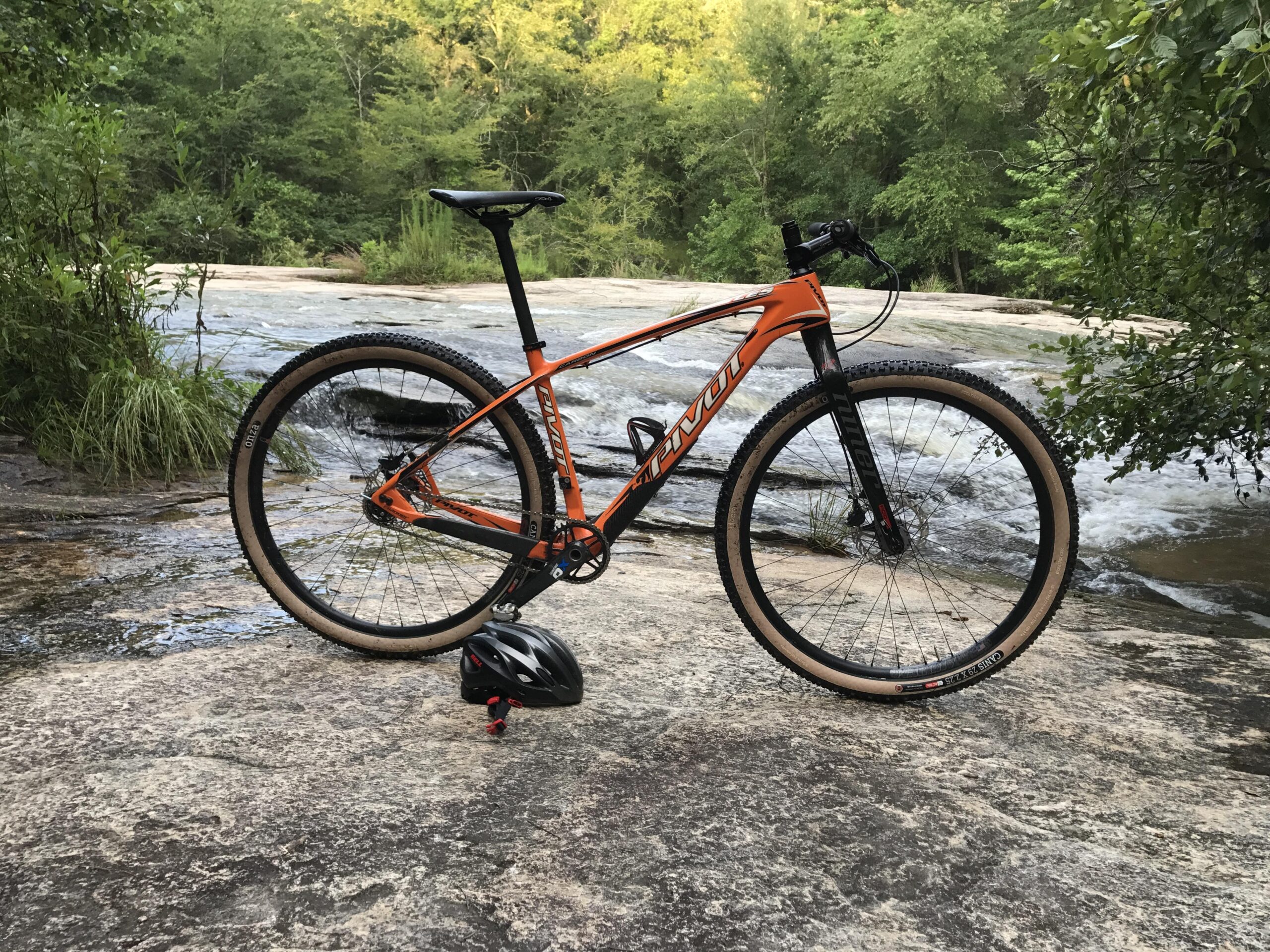 Pivot LES 29er: A bright orange mountain bike stands on a rocky surface near a flowing river. The background features lush green trees and foliage, creating a natural outdoor setting. A black helmet is placed on the ground next to the bike.