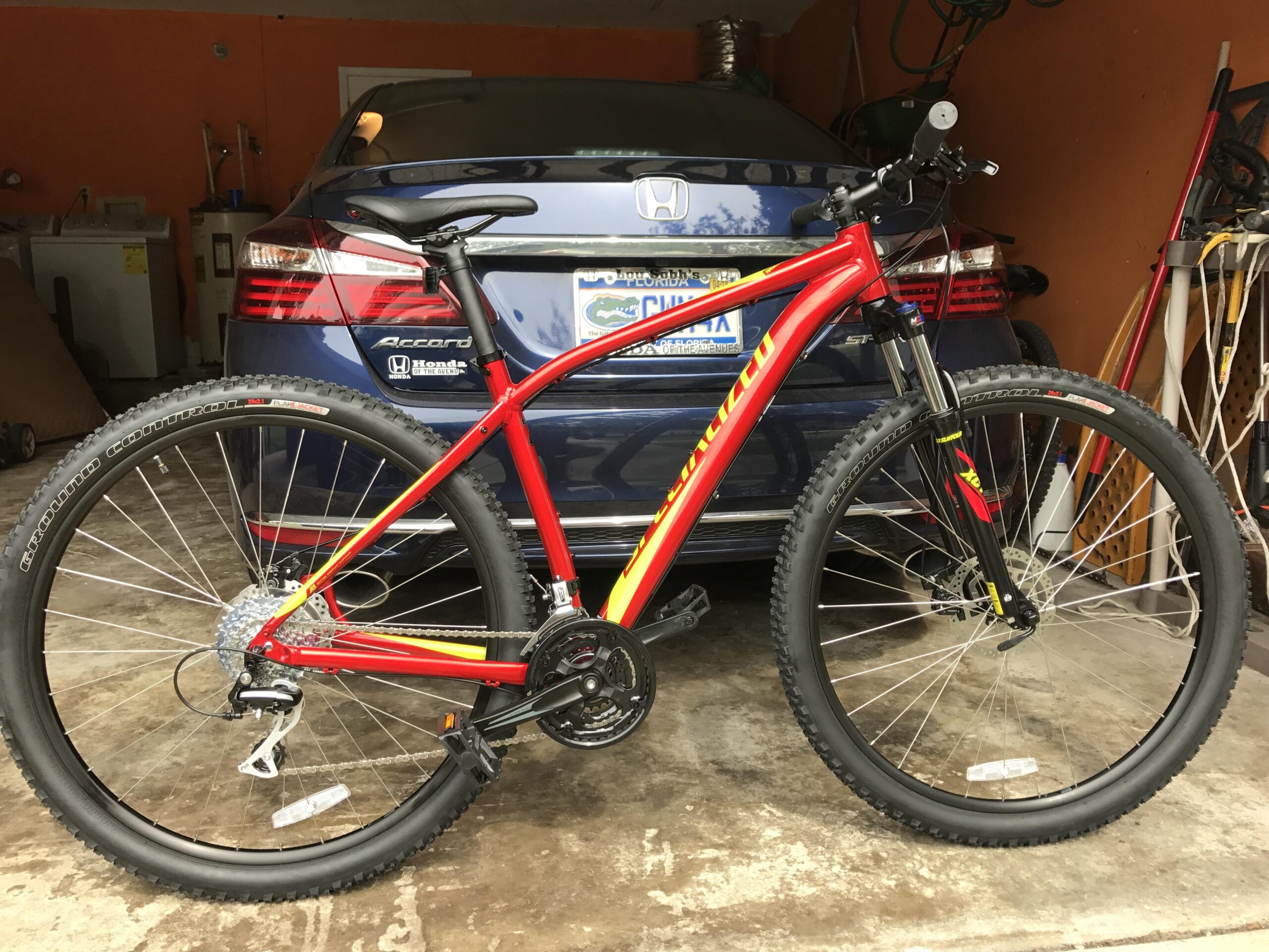 Specialized Rockhopper: A red mountain bike with black tires and front suspension is parked in a garage, with a blue Honda Accord visible in the background. The garage has orange walls and various tools and equipment around.