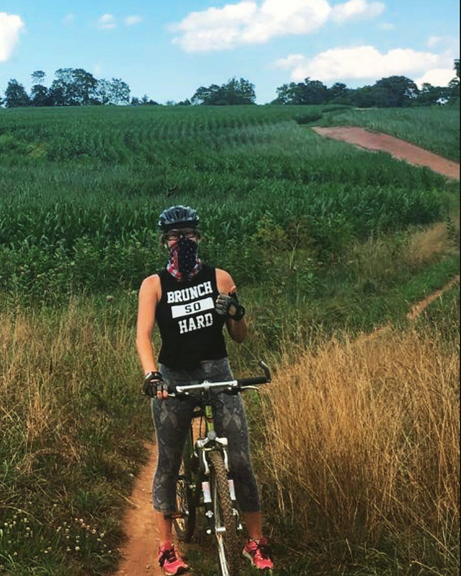 A cyclist wearing a black sleeveless shirt that says "BRUNCH SO HARD," a patterned face covering, and cycling gloves poses next to a mountain bike on a dirt trail surrounded by tall grass and green fields under a blue sky with scattered clouds. Schaeffer Farms mountain bike trail.