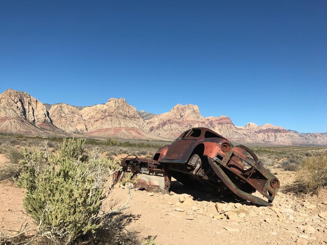 An abandoned, rusted car carcass sits on rocky terrain in the desert, with dramatic mountain peaks in the background under a clear blue sky. Sparse vegetation surrounds the vehicle, highlighting the arid landscape. Blue Diamond mountain bike trail.