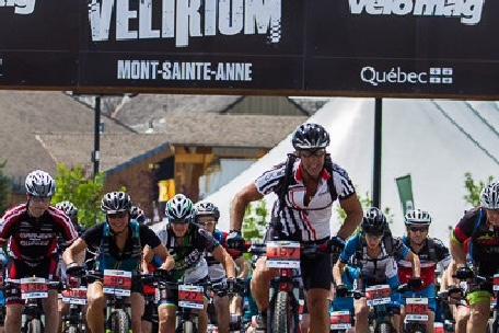 Felt Edict Nine 3: A group of mountain bikers in racing gear begins a competition under an archway banner that reads "VELIRIUM MONT-SAINTE-ANNE." The scene captures the excitement of the event as riders pedal aggressively, with various jerseys and numbers visible. The background includes trees and a tent.