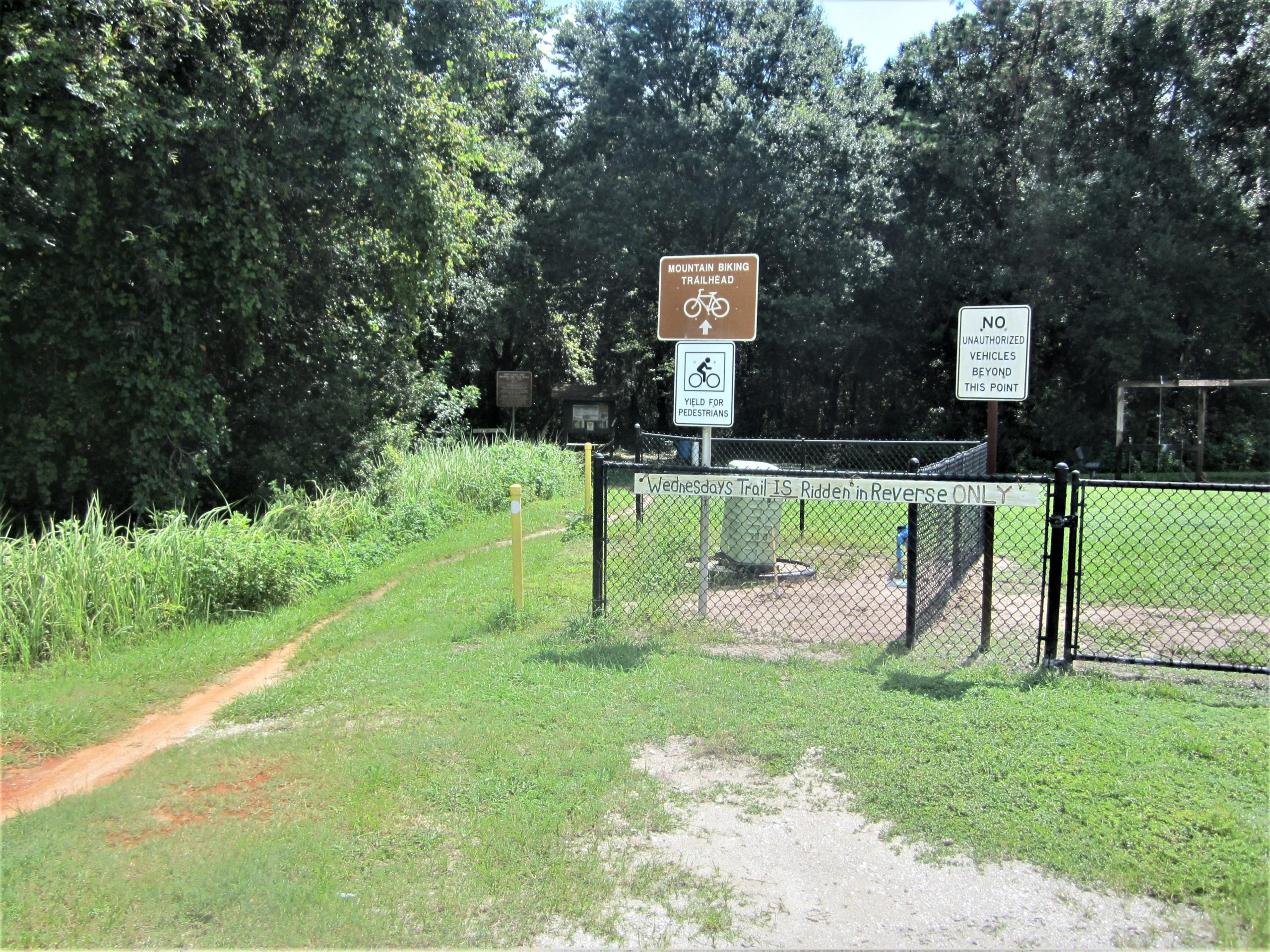 Mountain biking trailhead entrance featuring signs for trail usage, including "Wednesday's Trail IS Ridden in Reverse ONLY," and instructions for yielding to pedestrians. A fenced area with a path leading into a wooded area is visible, surrounded by tall grass and trees. Loyce E. Harpe Park mountain bike trail.