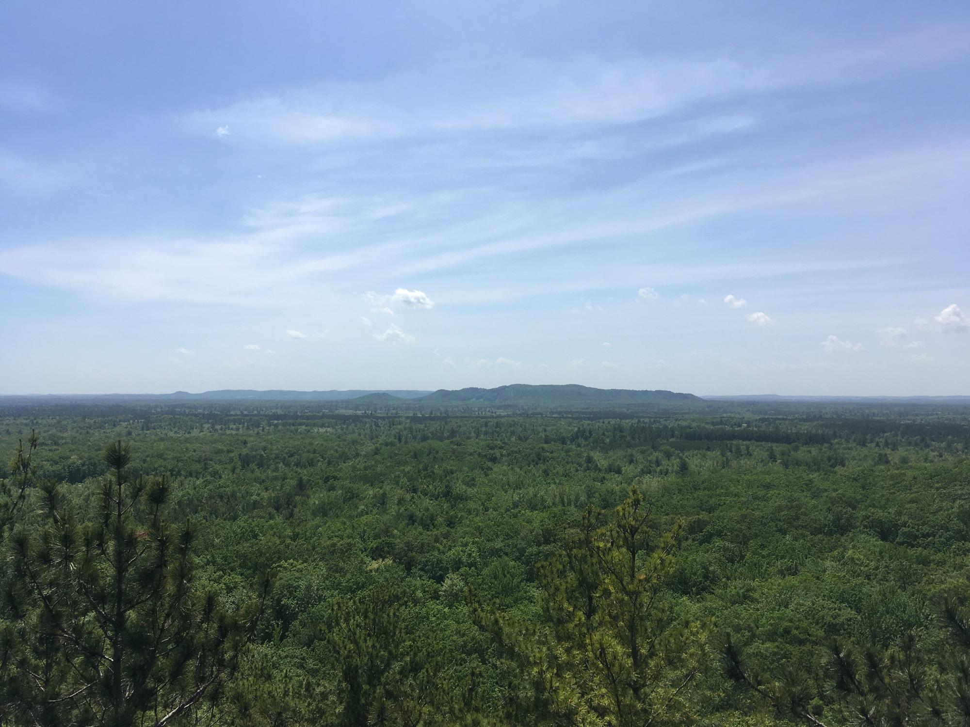 A panoramic view of a lush green landscape under a clear blue sky, featuring rolling hills in the distance and a dense forest covering the foreground. The scene is lit by bright sunlight, with a few wispy clouds scattered across the sky. Levis Mounds mountain bike trail.