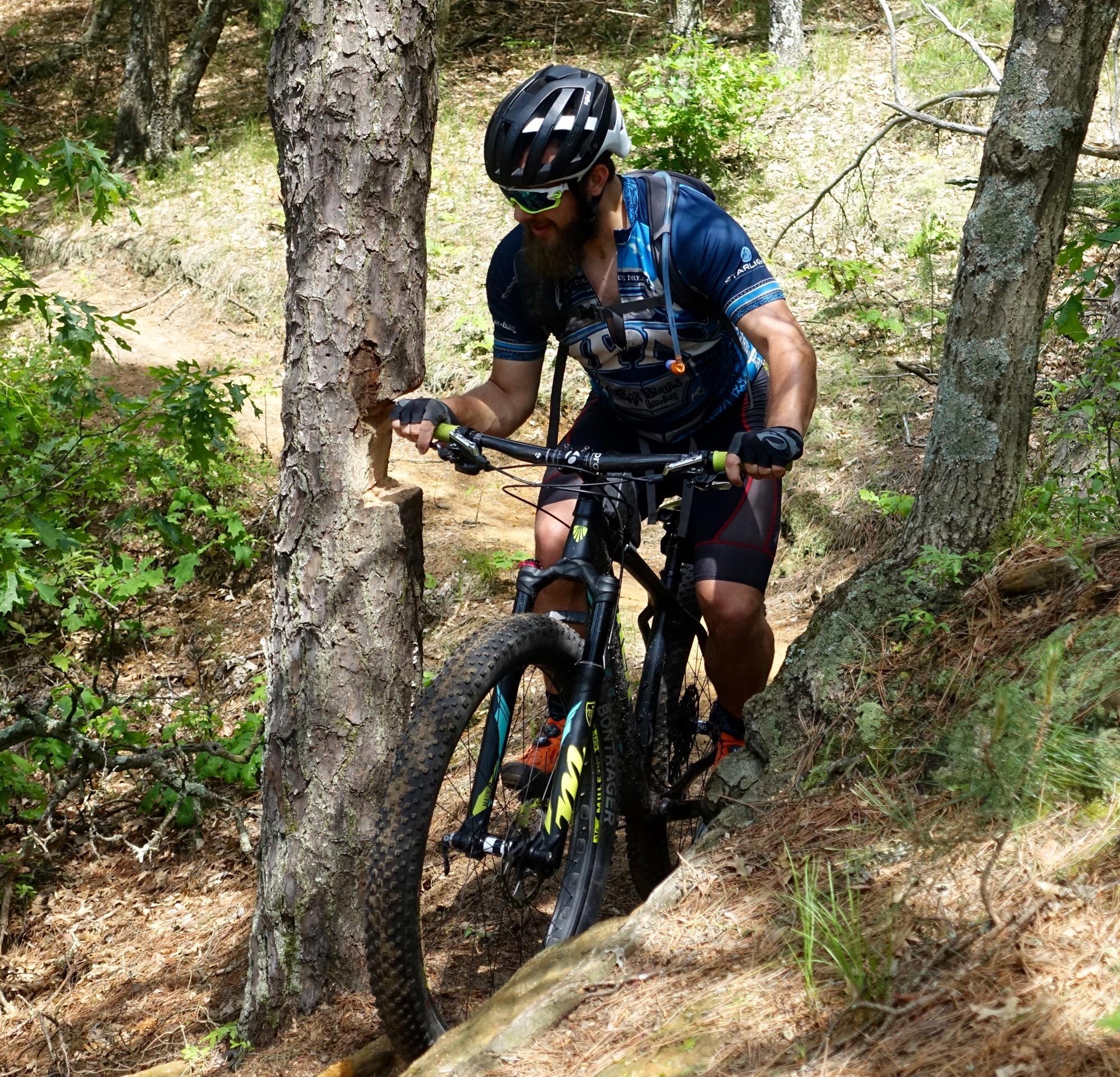 A mountain biker navigating a narrow trail, climbing beside a tree with visible bark damage. The biker, wearing a helmet and sunglasses, is focused on maneuvering the bike over rough terrain, surrounded by greenery and dirt pathways. Levis Mounds mountain bike trail.