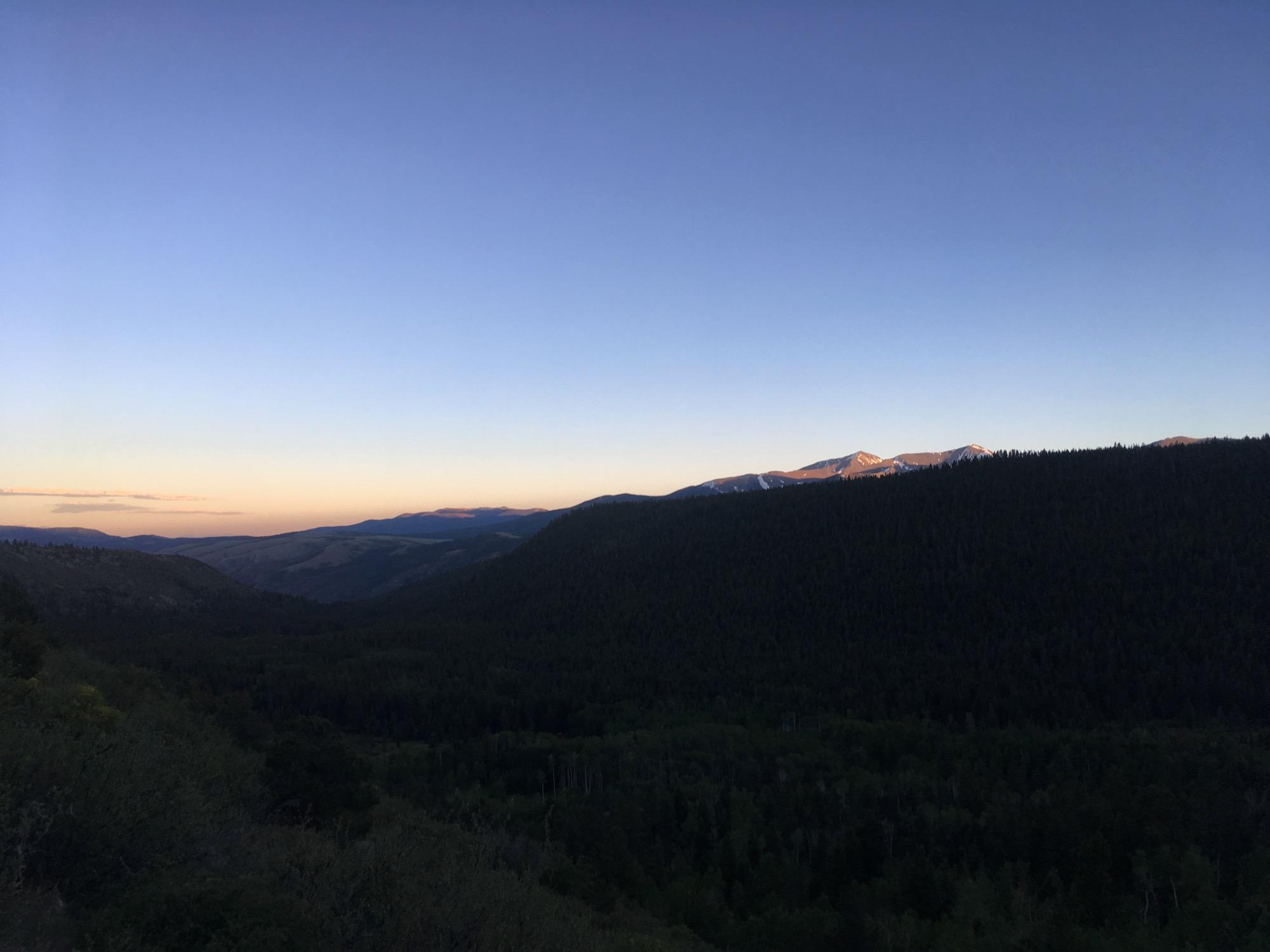A tranquil mountain landscape at dusk, featuring rolling hills and dense evergreen forests with a backdrop of snow-capped peaks. The sky transitions from light blue to soft hues of orange and pink, indicating sunset. Colorado Trail: Hwy 50 to Mt. Shavano Trailhead mountain bike trail.