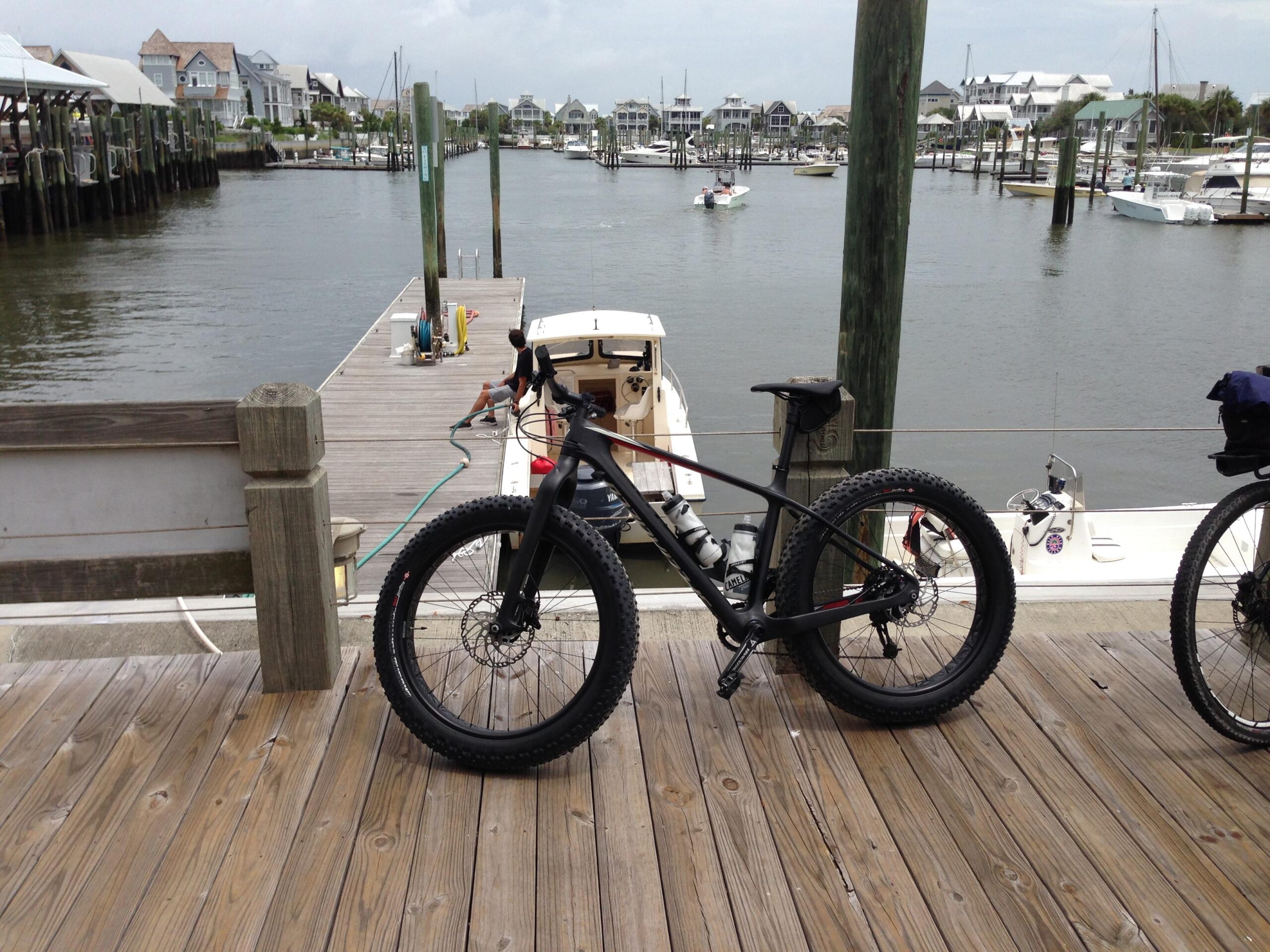 Specialized Fatboy Carbon Comp: A fat tire bike parked on a wooden dock overlooking a calm waterway, with boats and a marina in the background. The scene is set in a coastal area with houses visible in the distance, and a person is seen working on a boat.