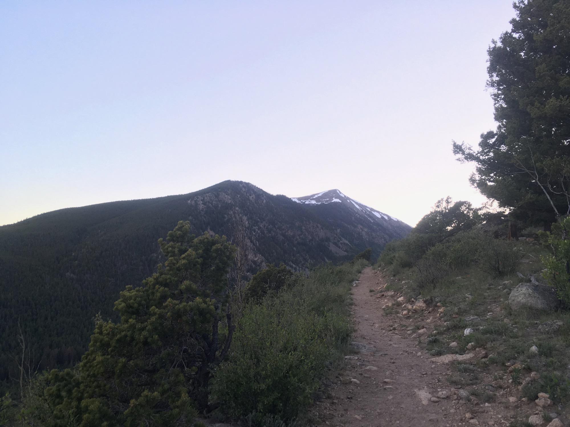 A winding dirt path leads through a mountainous landscape, flanked by green bushes and trees, with snow-capped peaks in the distance under a clear sky. Colorado Trail: Hwy 50 to Mt. Shavano Trailhead mountain bike trail.