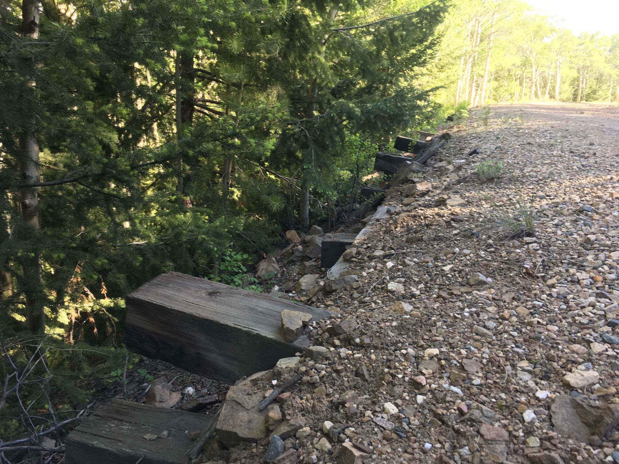 A dirt path alongside a wooded area, featuring exposed wooden beams and scattered rocks. Lush green trees are visible on both sides of the path, with sunlight filtering through the foliage. Marshall Pass Road / #200 / #203 / #243 mountain bike trail.