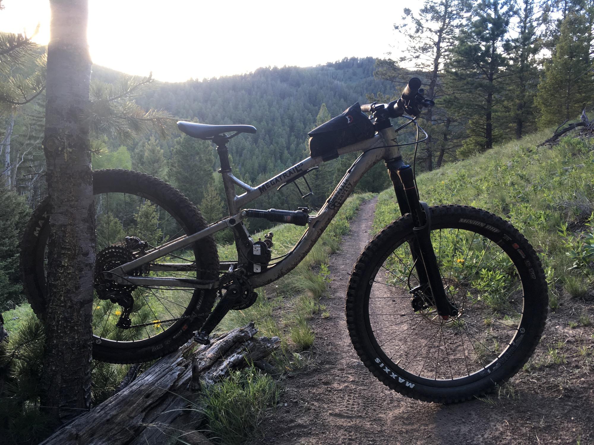 A mountain bike resting against a tree on a dirt trail, surrounded by greenery and rolling hills under a soft, golden light. The bike features thick tires and a suspension system, suggesting it is designed for rugged terrain. The background shows dense forests and a sloping landscape. Rainbow Trail: Silver Creek to Hwy 285 mountain bike trail.