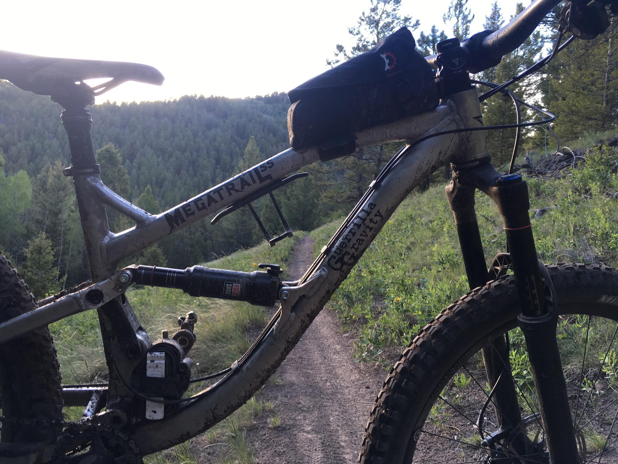A close-up view of a mountain bike's frame with the words "MEGATRAILS" and "Guerilla Gravity" visible, showcasing a dirty, well-used design. The bike is positioned on a dirt trail surrounded by lush greenery and trees, capturing a scenic outdoor environment in the late afternoon light. Rainbow Trail: Silver Creek to Hwy 285 mountain bike trail.