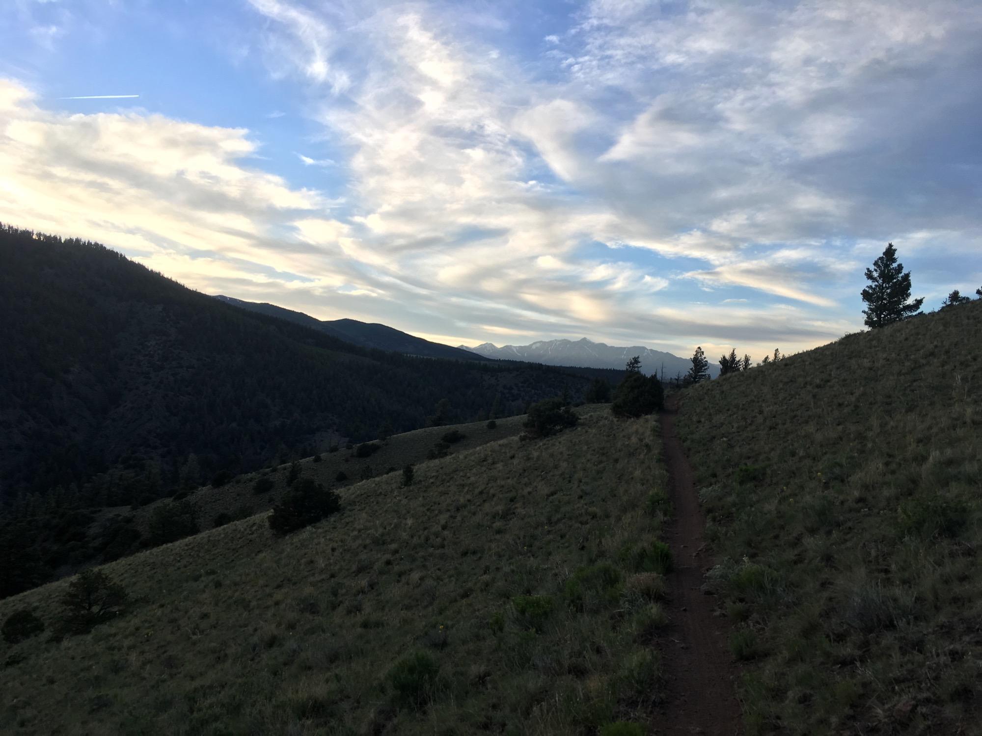 A scenic view of rolling hills under a partly cloudy sky, featuring a winding dirt trail leading through grassy terrain. In the distance, mountain peaks are visible, framed by lush trees on the slopes. Rainbow Trail: Silver Creek to Hwy 285 mountain bike trail.