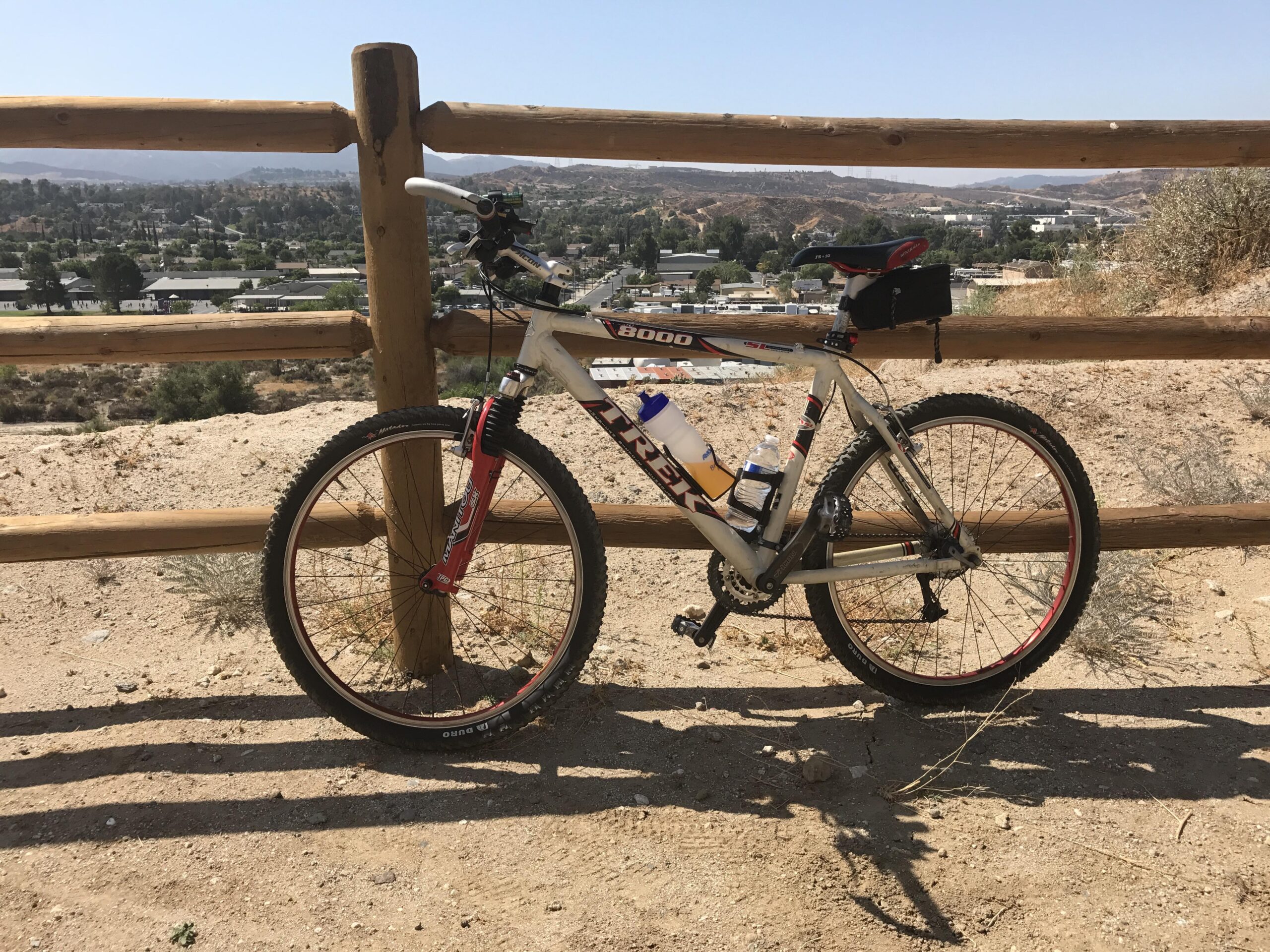 Trek 8000: A mountain bike leaning against a wooden fence with a scenic view of a hilly landscape and distant buildings. The bike has a water bottle holder, and the ground is sandy with sparse vegetation.