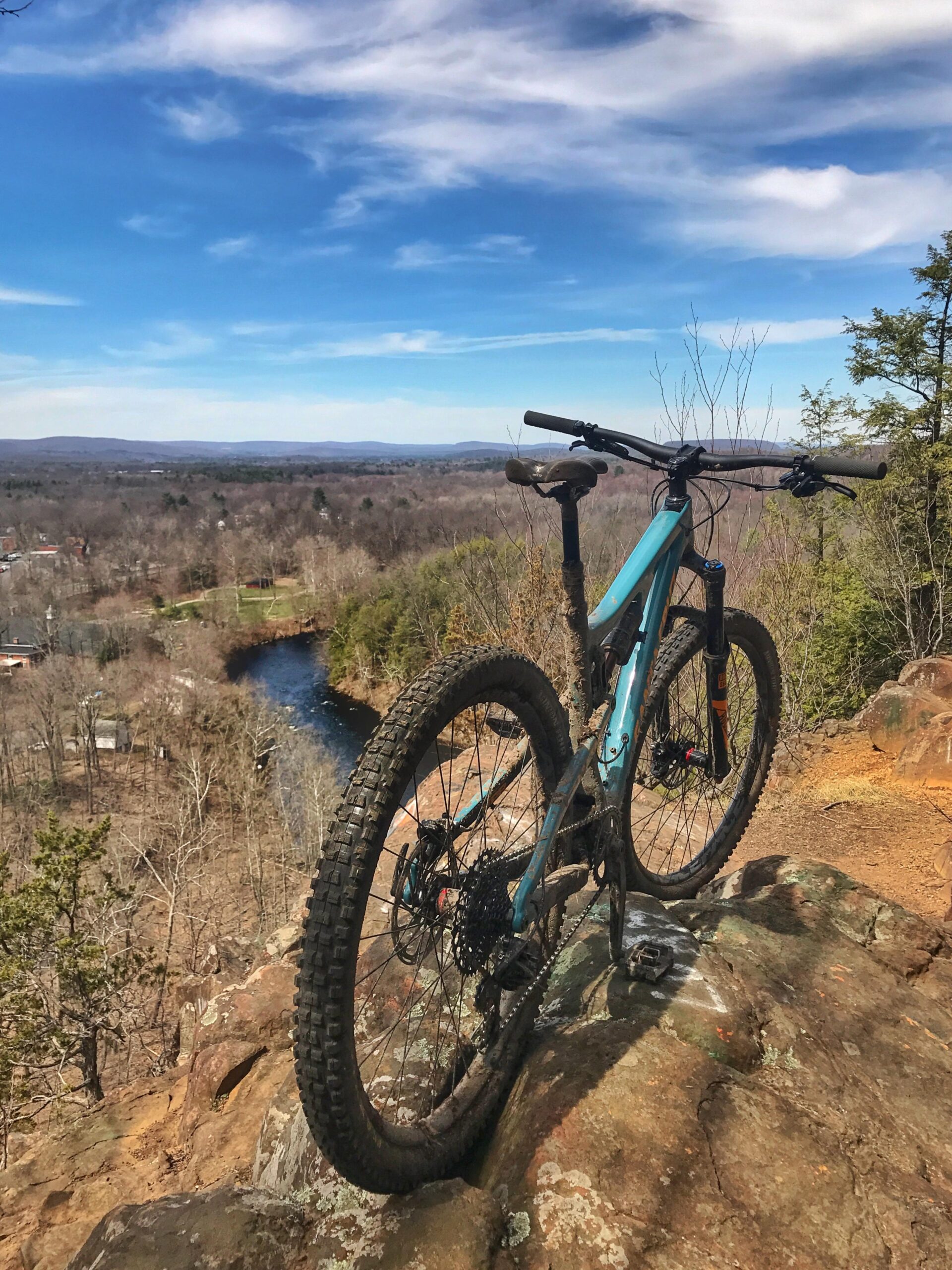 Santa Cruz Bronson: A mountain bike rests on a rocky ledge overlooking a river and wooded landscape under a bright blue sky with wispy clouds. The scene captures the beauty of nature and outdoor adventure, highlighting the bike as a focal point.