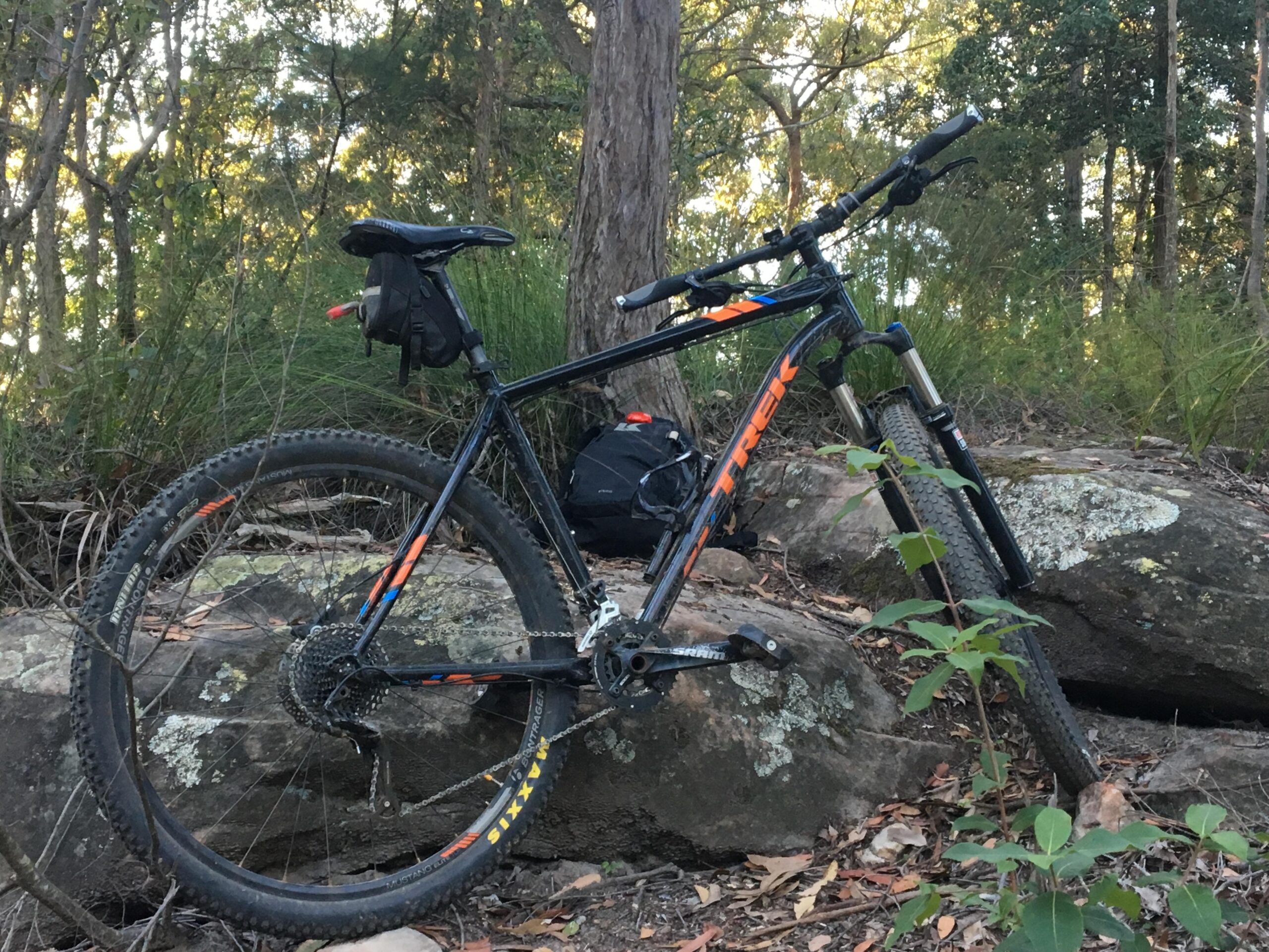 Trek Trek x caliber 8: A black mountain bike with orange accents rests against a large rock in a forested area. Surrounding the bike are trees and shrubs, creating a natural outdoor setting. A small backpack is positioned on the ground nearby.