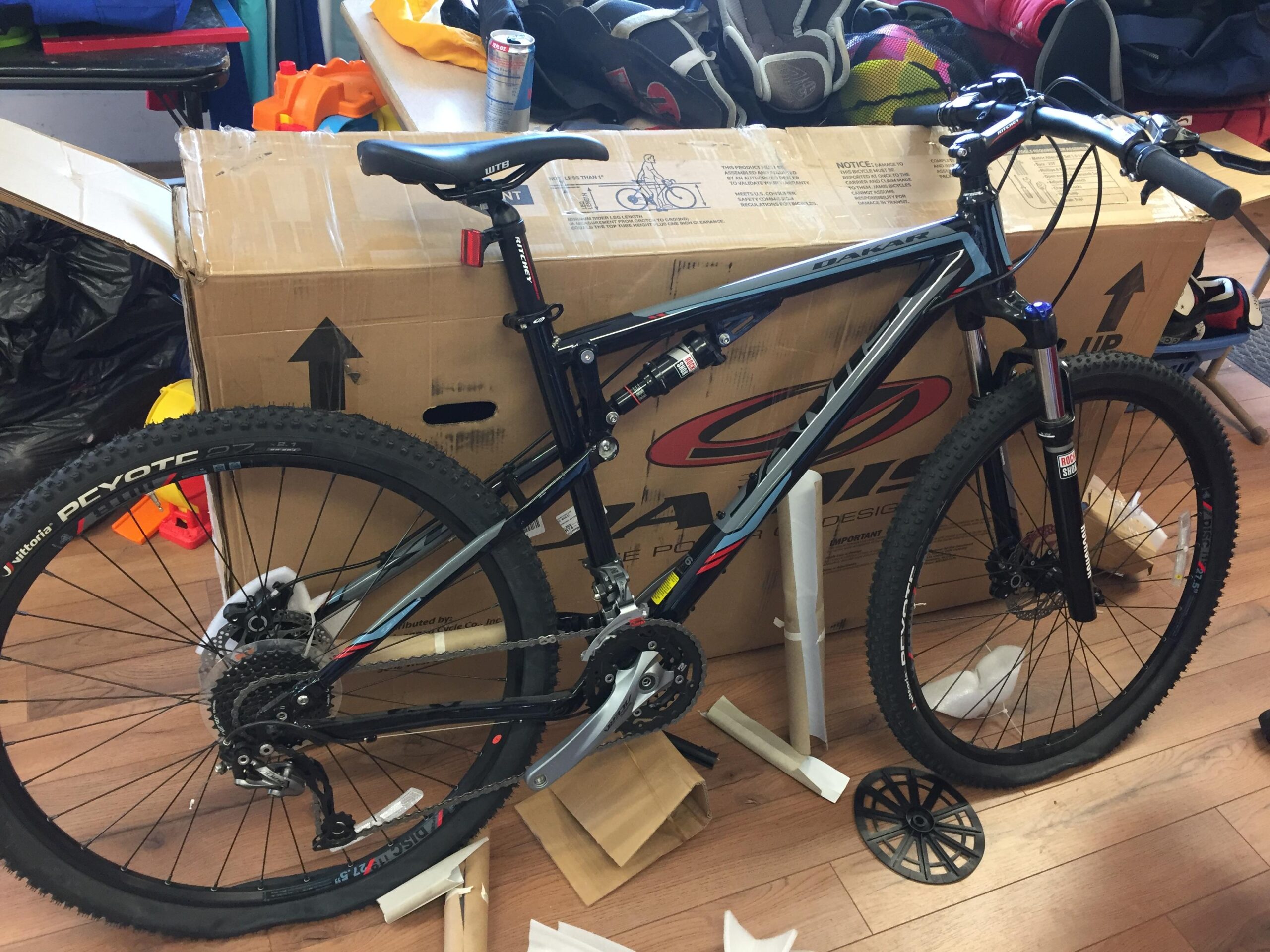 Jamis Dakar XC: A black and blue mountain bike is standing next to an open cardboard box. The bike features thick tires and a sturdy frame, with various gears and components visible. Behind the bike, there are colorful toys and bags, indicating a cluttered indoor space. Some packaging materials are scattered on the floor.