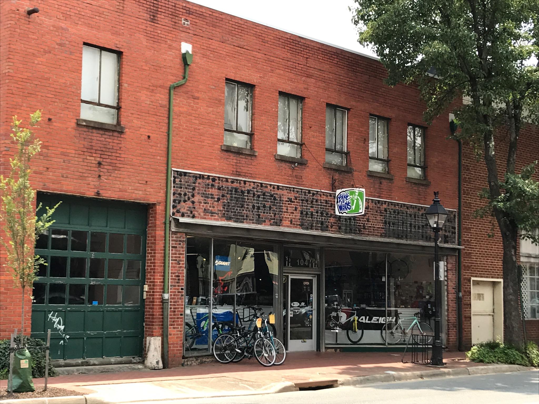Alt tag: A brick building with large windows and a green garage door, featuring a bicycle shop front displaying various bicycles and accessories. Trees and a lamp post are visible in front of the building.