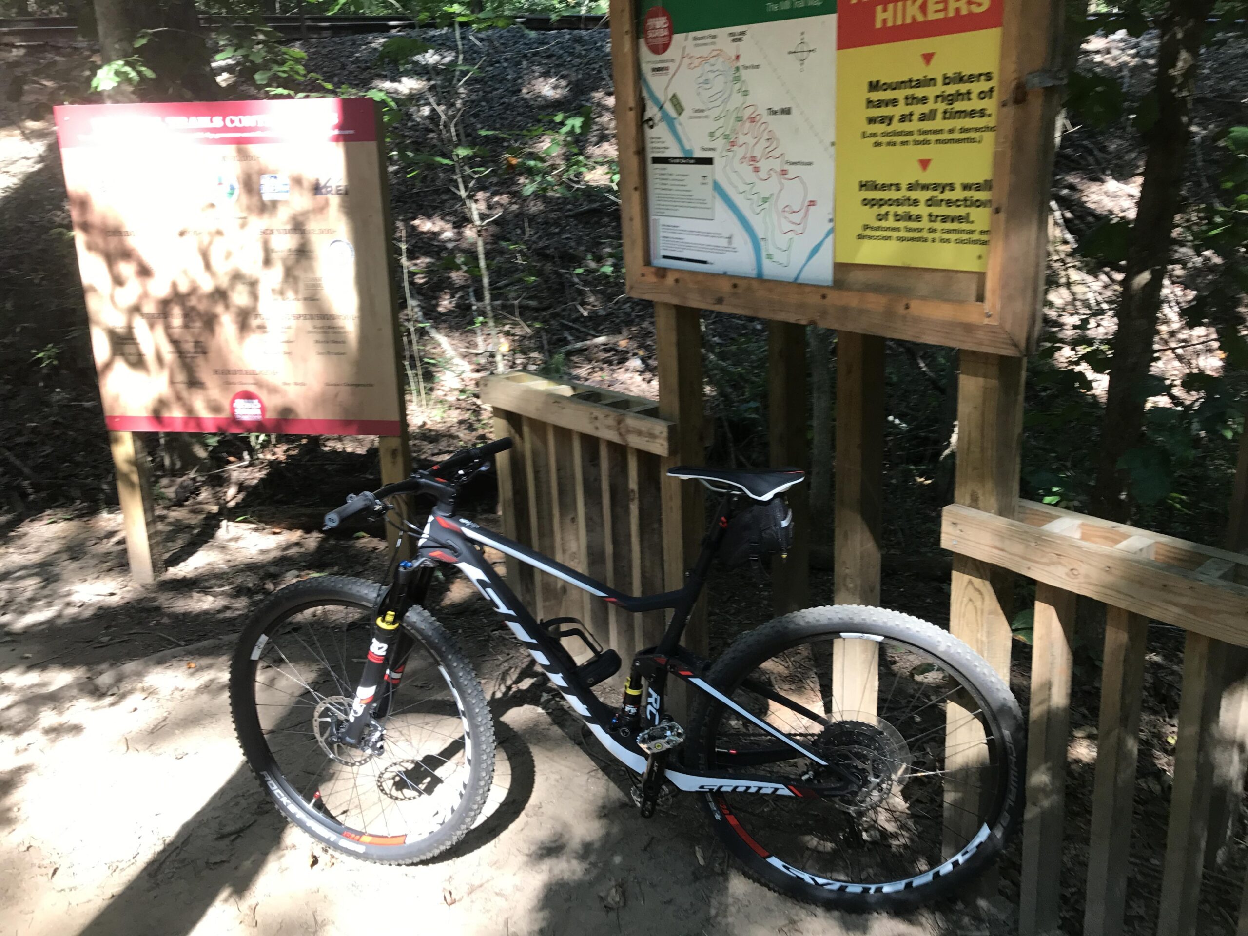 Scott 900RC 29er: A black and red mountain bike is leaned against a wooden structure near a trail, with a signboard visible in the background that provides information for hikers and mountain bikers. The area is wooded, with sunlight filtering through the trees.