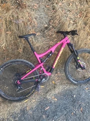 Santa Cruz Bronson: A vibrant pink mountain bike resting against a dry grass backdrop, showcasing its sturdy frame and rugged tires, positioned on a gravel surface.