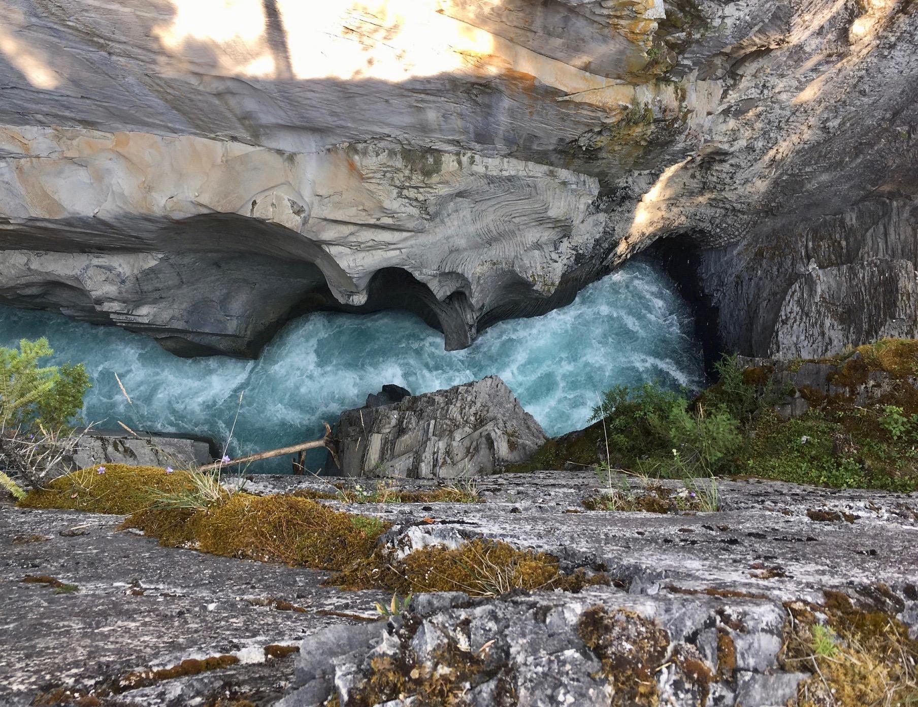 Aerial view of a turquoise river rushing through a rocky gorge, surrounded by rugged stone cliffs and patches of green vegetation. The water creates frothy waves as it flows swiftly over boulders and along the rocky edges. Sunlight filters through, casting patterns on the rock surface. Nipika Mountain Resort mountain bike trail.