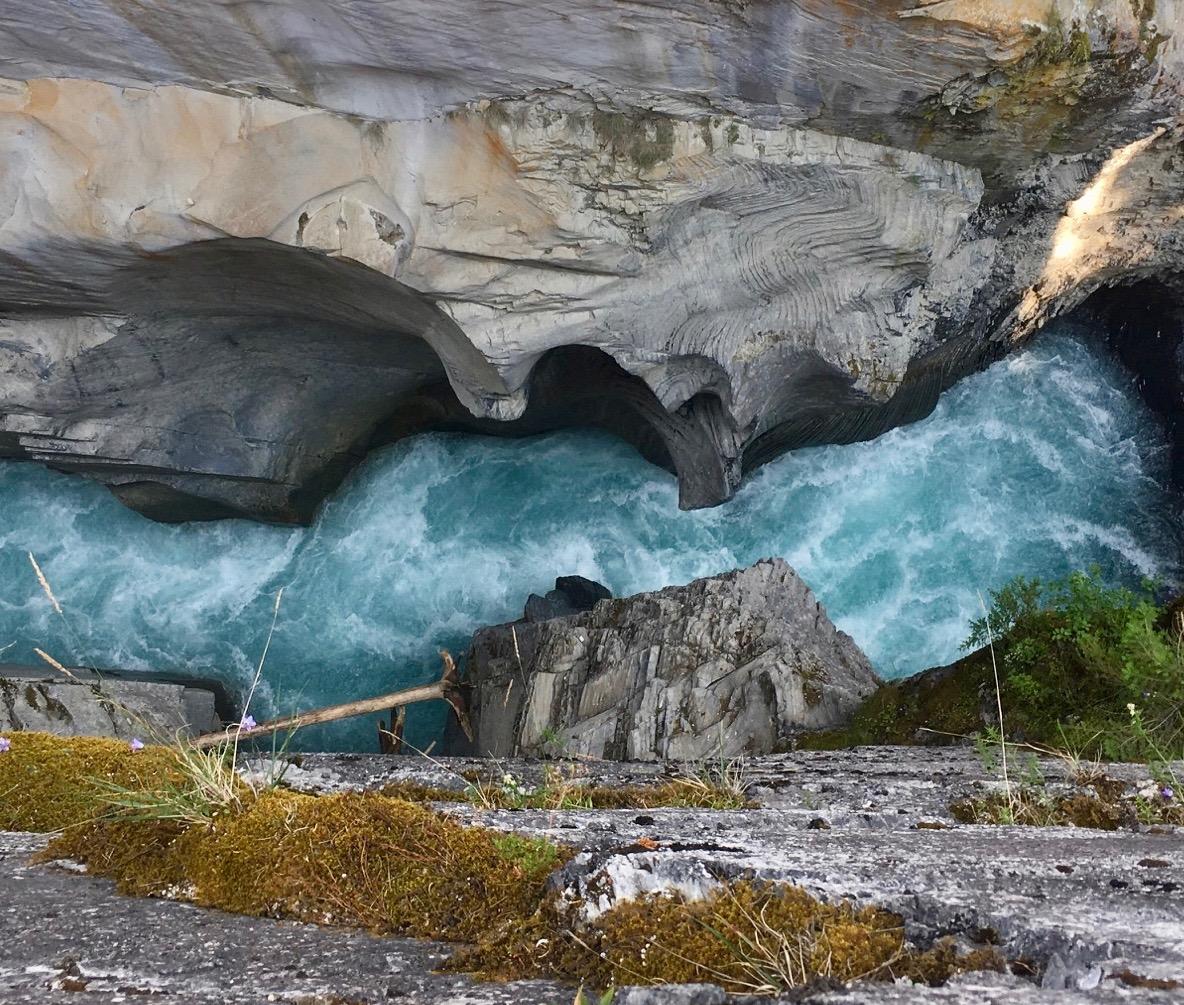 Aerial view of a rushing turquoise river flowing through a rocky gorge, surrounded by textured stone walls and patches of green moss. A fallen branch lies across the rocky ledge, adding a natural element to the scene. Nipika Mountain Resort mountain bike trail.
