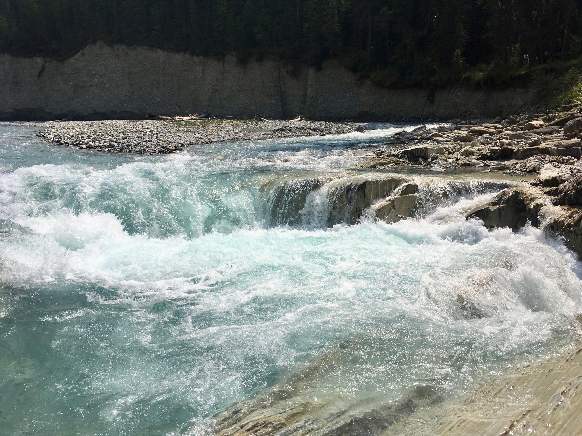 A gentle river cascading over rocky ledges, creating frothy white water as it flows. The surrounding landscape features a mix of smooth stones and a backdrop of trees and cliffs, with clear blue water reflecting sunlight. Nipika Mountain Resort mountain bike trail.