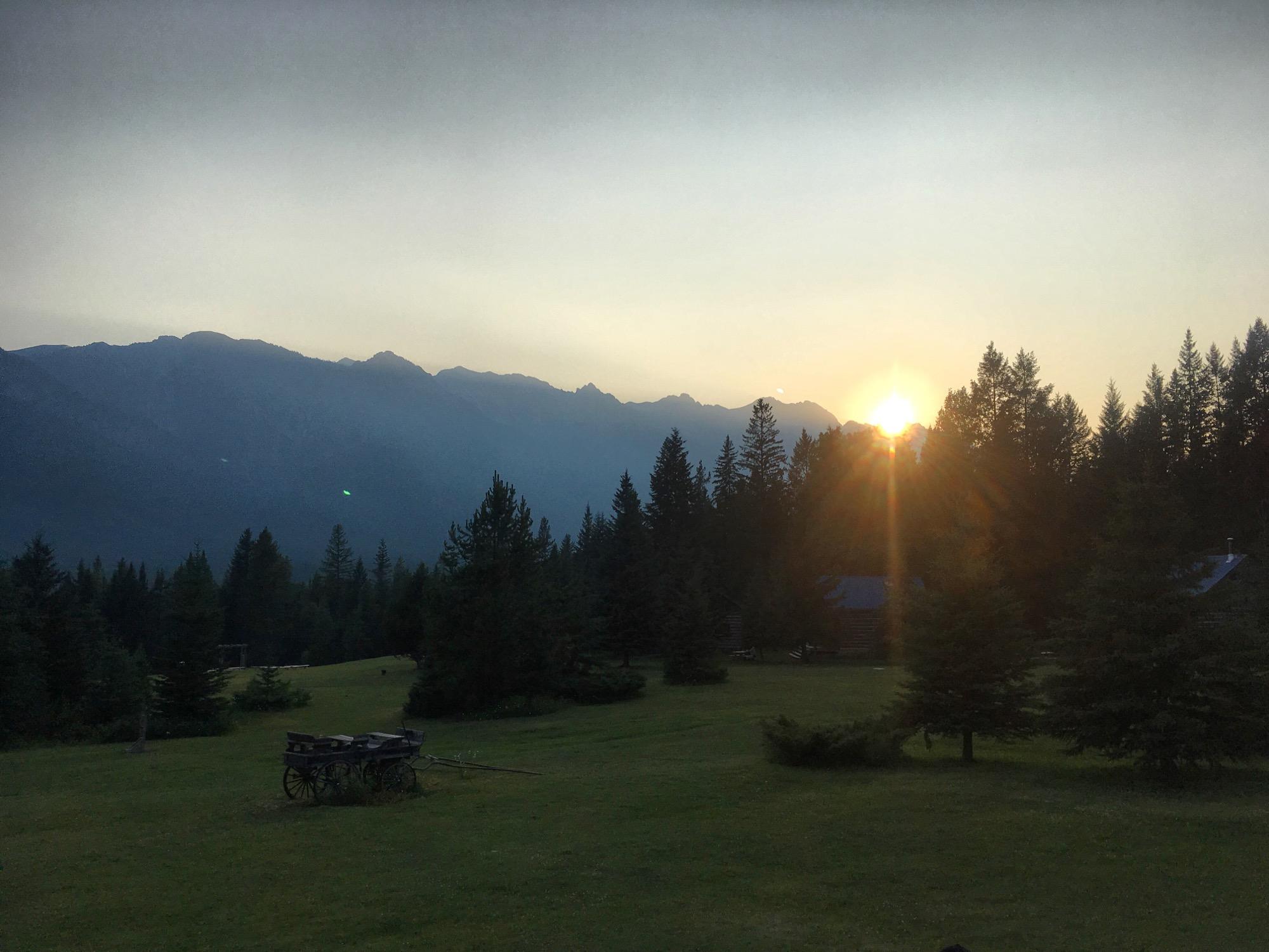 Sunset over a mountainous landscape, with the sun partially hidden behind the peaks. The foreground features a grassy field dotted with trees and a rustic wooden cart, creating a serene natural scene. The sky is hazy, transitioning from light to darker shades as evening approaches. Nipika Mountain Resort mountain bike trail.