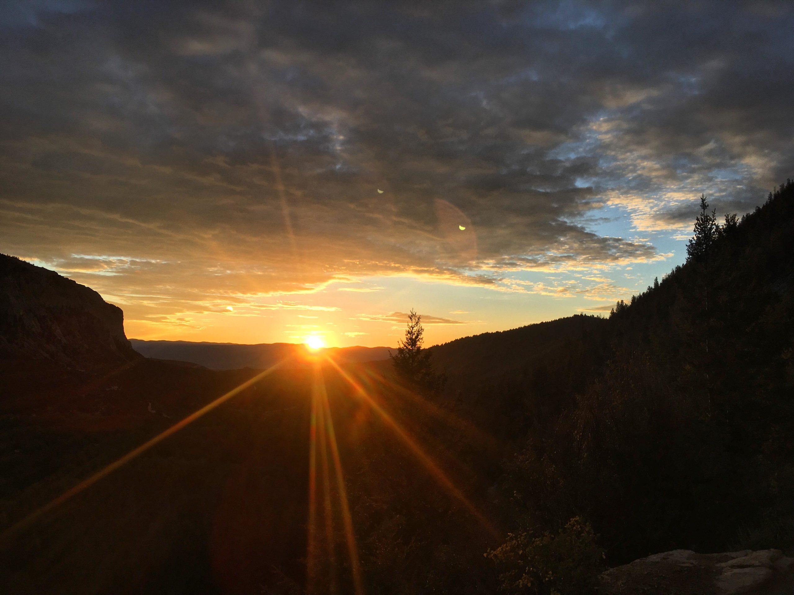 A breathtaking sunset over a mountainous landscape, with vibrant orange and yellow hues illuminating the sky. Silhouettes of trees and rocky cliffs frame the scene, while rays of sunlight create a dramatic effect as the sun dips below the horizon. The sky features a mix of clouds, adding depth and texture to the tranquil setting. Narrow Gauge Trail mountain bike trail.