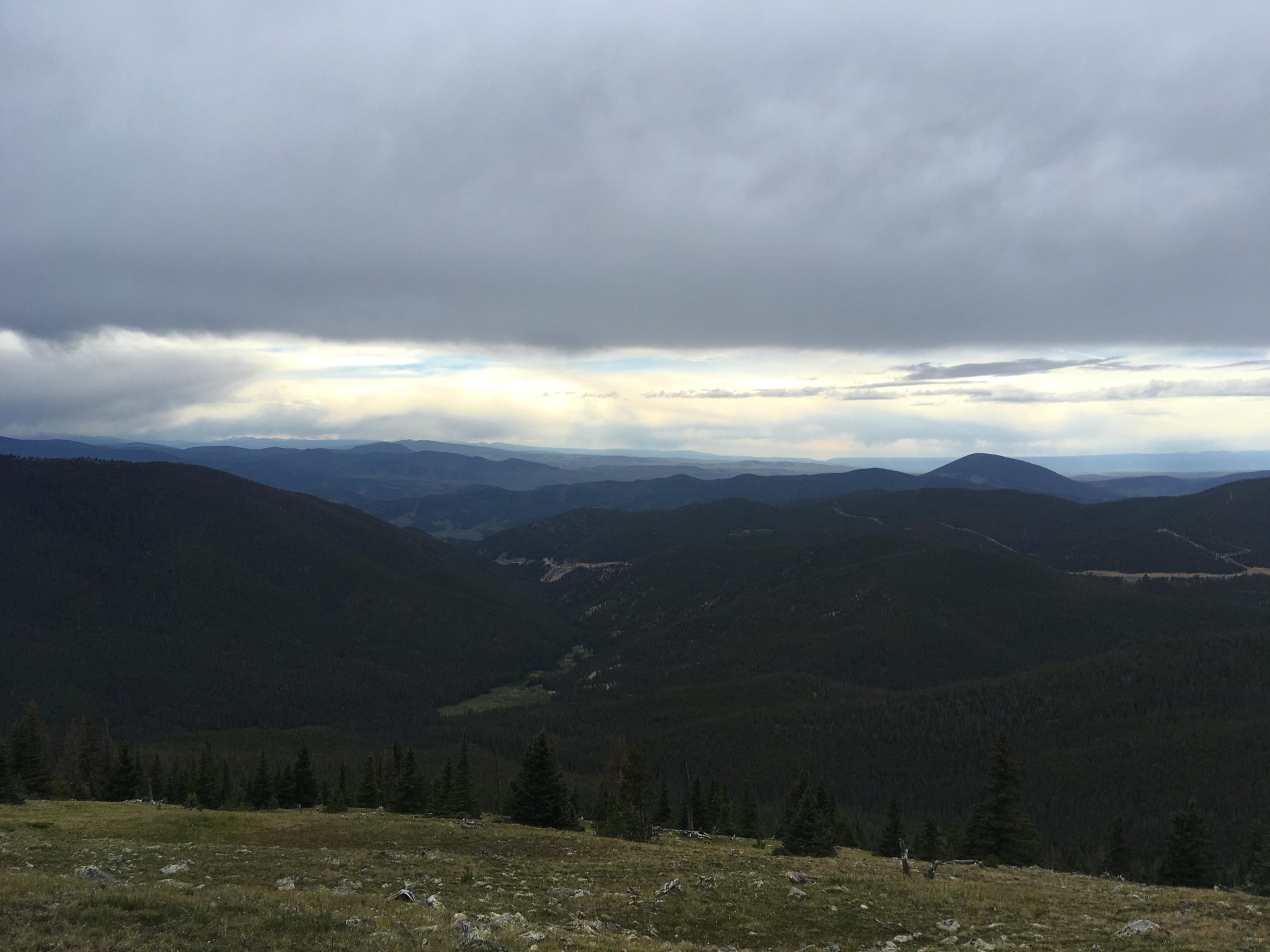 A panoramic view of rolling green mountains under a cloudy sky, with distant hills visible in the background, capturing the tranquility of nature. Monarch Crest Trail mountain bike trail.