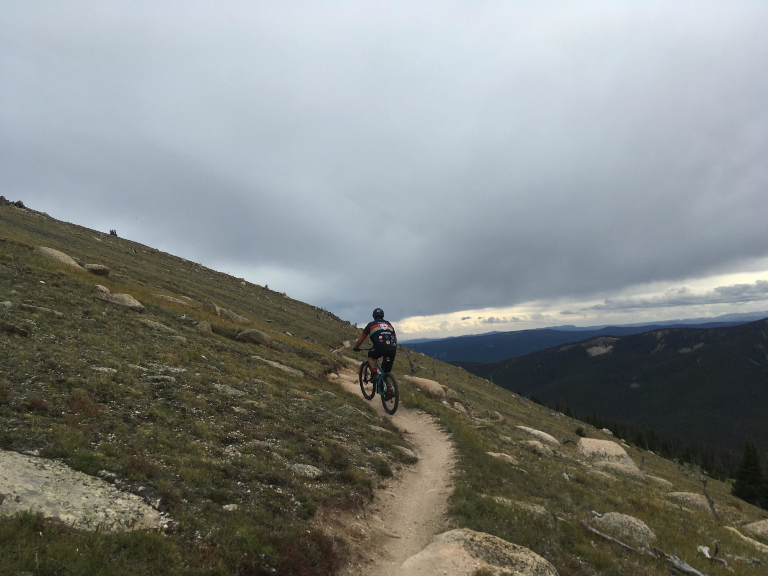 A mountain biker riding along a winding dirt trail on a grassy hillside, with rocky terrain and cloudy skies in the background, showcasing a panoramic view of distant mountains and valleys. Monarch Crest Trail mountain bike trail.
