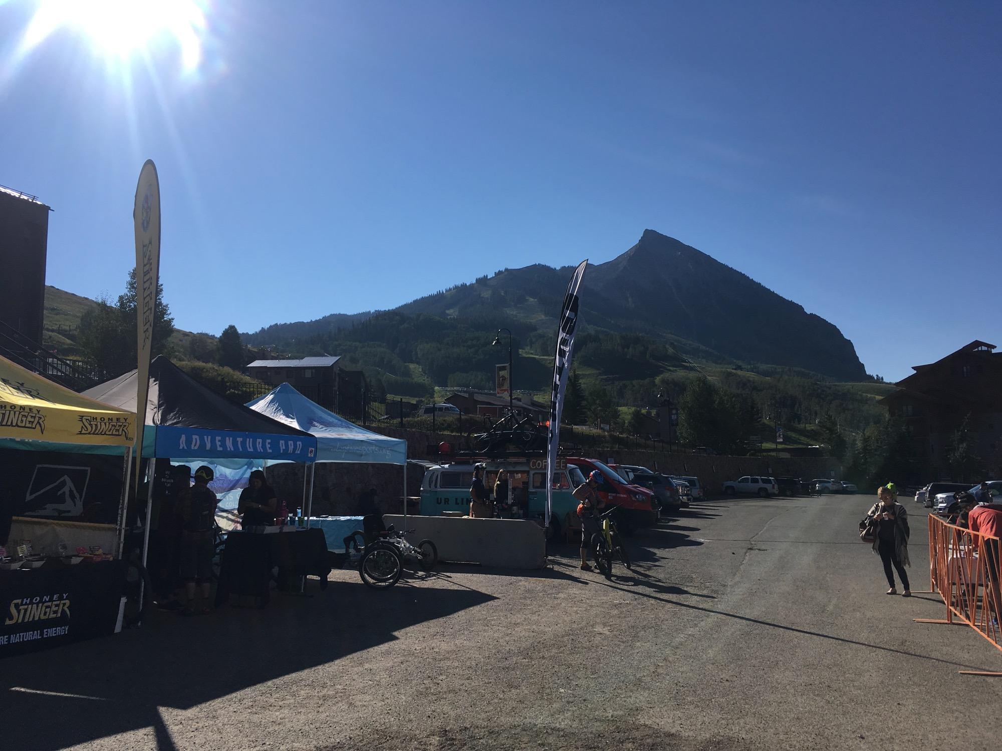 A bright, sunny day at a mountain event with tents displaying different brands, including Honey Stinger and Adventure Pod. In the background, a prominent mountain peak is visible, and parked vehicles line the gravel road. People are gathered around the tents, engaging in activities, while race barriers are set up nearby. Evolution Bike Park at Crested Butte Mountain Resort mountain bike trail.