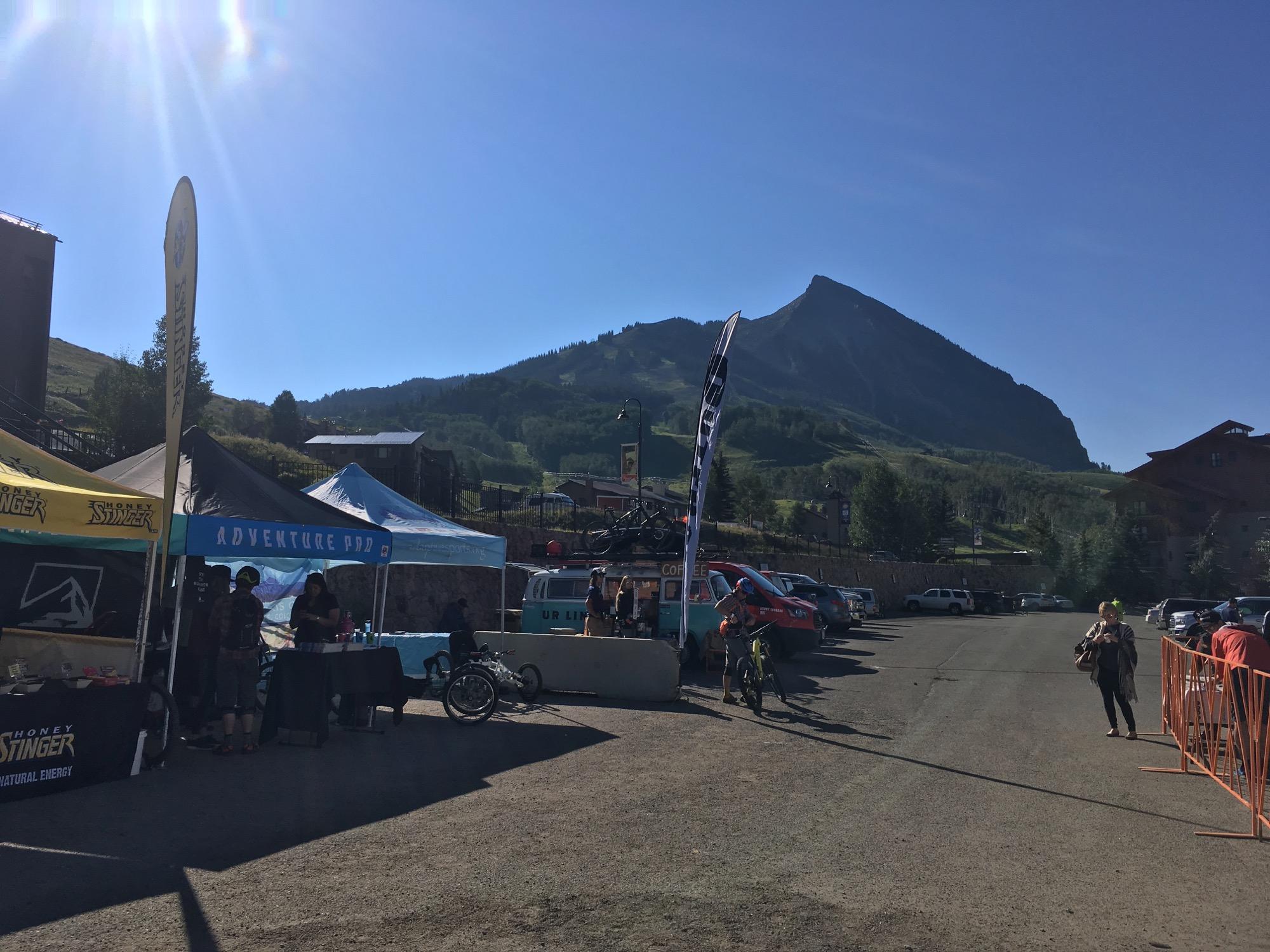 A sunny day at an outdoor event featuring vendor tents, including "Adventure Pod" and "Honey Stinger," with people milling around. In the background, a mountain rises under a clear blue sky. Several parked cars are visible along the gravel road, creating a lively atmosphere perfect for outdoor activities. Evolution Bike Park at Crested Butte Mountain Resort mountain bike trail.
