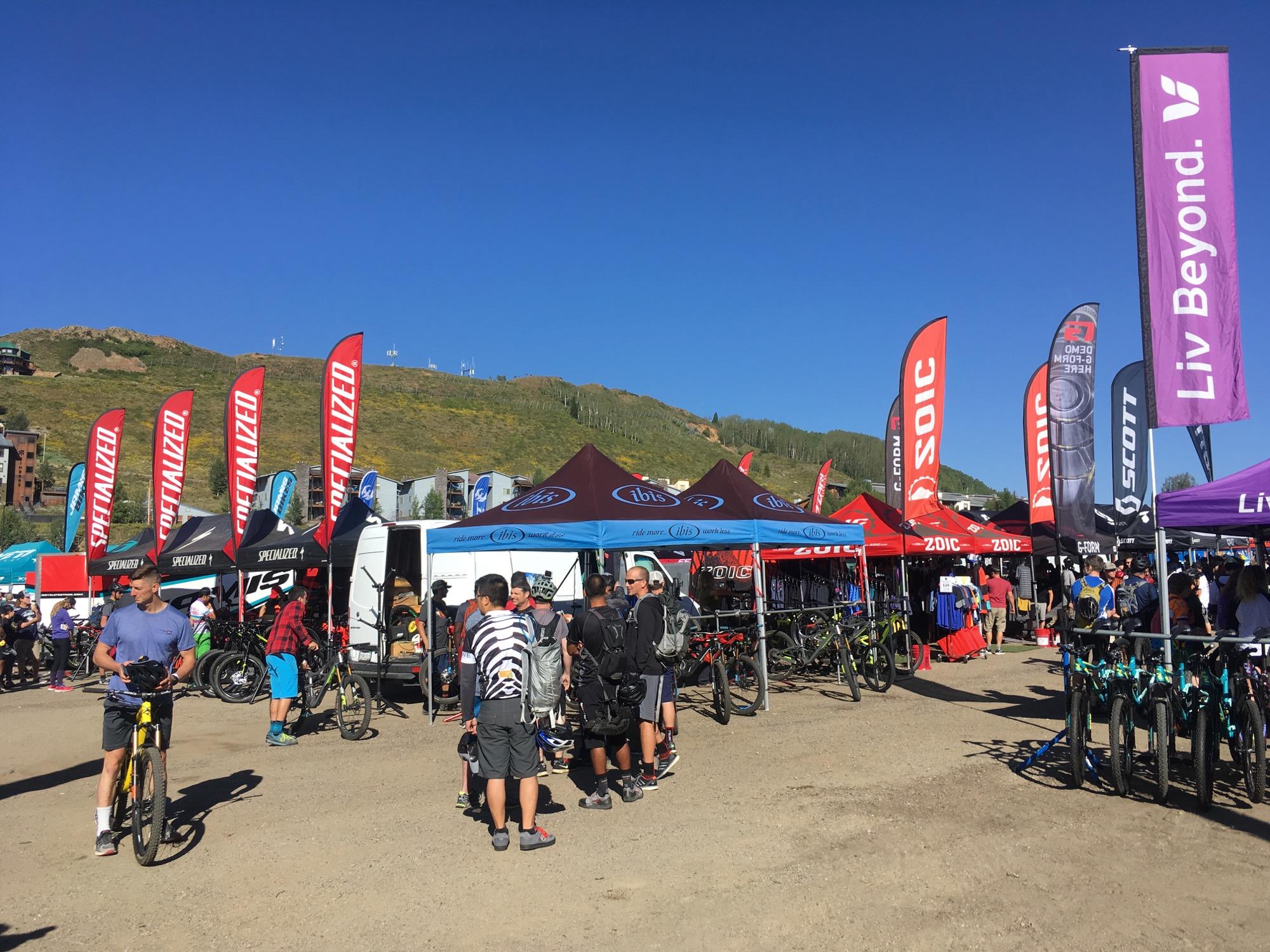 A vibrant outdoor scene at a biking festival, featuring various tents and flags from cycling brands like Specialized and Liv. People are gathered around, interacting and exploring numerous bikes displayed. The backdrop includes a clear blue sky and a hillside with greenery, indicating a sunny day suitable for outdoor activities. Evolution Bike Park at Crested Butte Mountain Resort mountain bike trail.