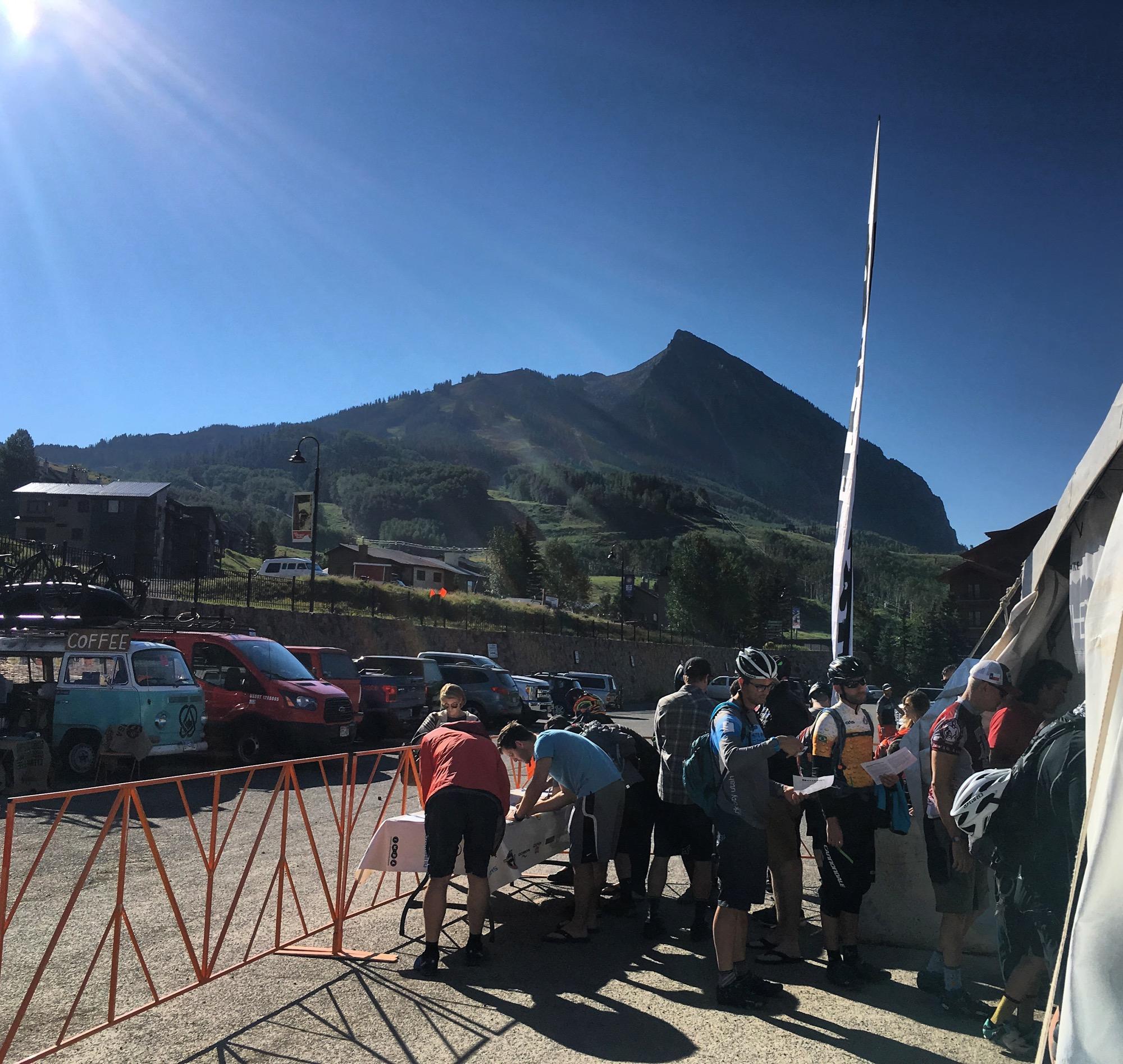 A sunny outdoor scene at a mountain location, featuring a group of cyclists gathered near a registration area. In the background, the prominent peak of a mountain rises against a clear blue sky. Several vehicles are parked nearby, including a food truck with a "COFFEE" sign. Cyclists are interacting by a table, some checking in for an event. Evolution Bike Park at Crested Butte Mountain Resort mountain bike trail.