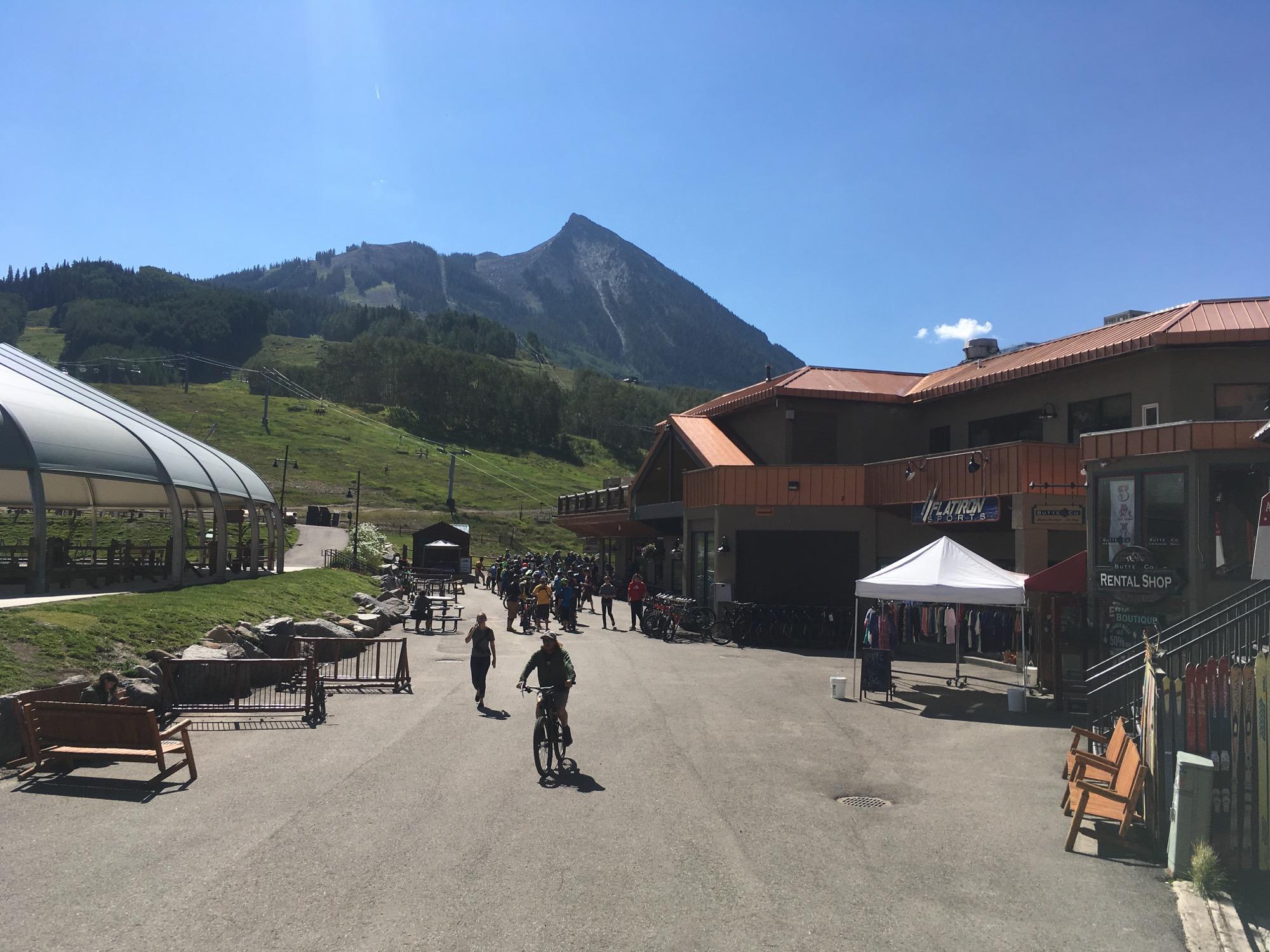 A sunny day at a mountain resort with a clear blue sky. In the foreground, a paved path leads from a rental shop with outdoor seating to a bustling area where people gather, some on bicycles. To the left, a semi-enclosed dining area and benches are visible, while a ski lift can be seen on the hillside behind the resort, leading up to a mountainous backdrop. Evolution Bike Park at Crested Butte Mountain Resort mountain bike trail.