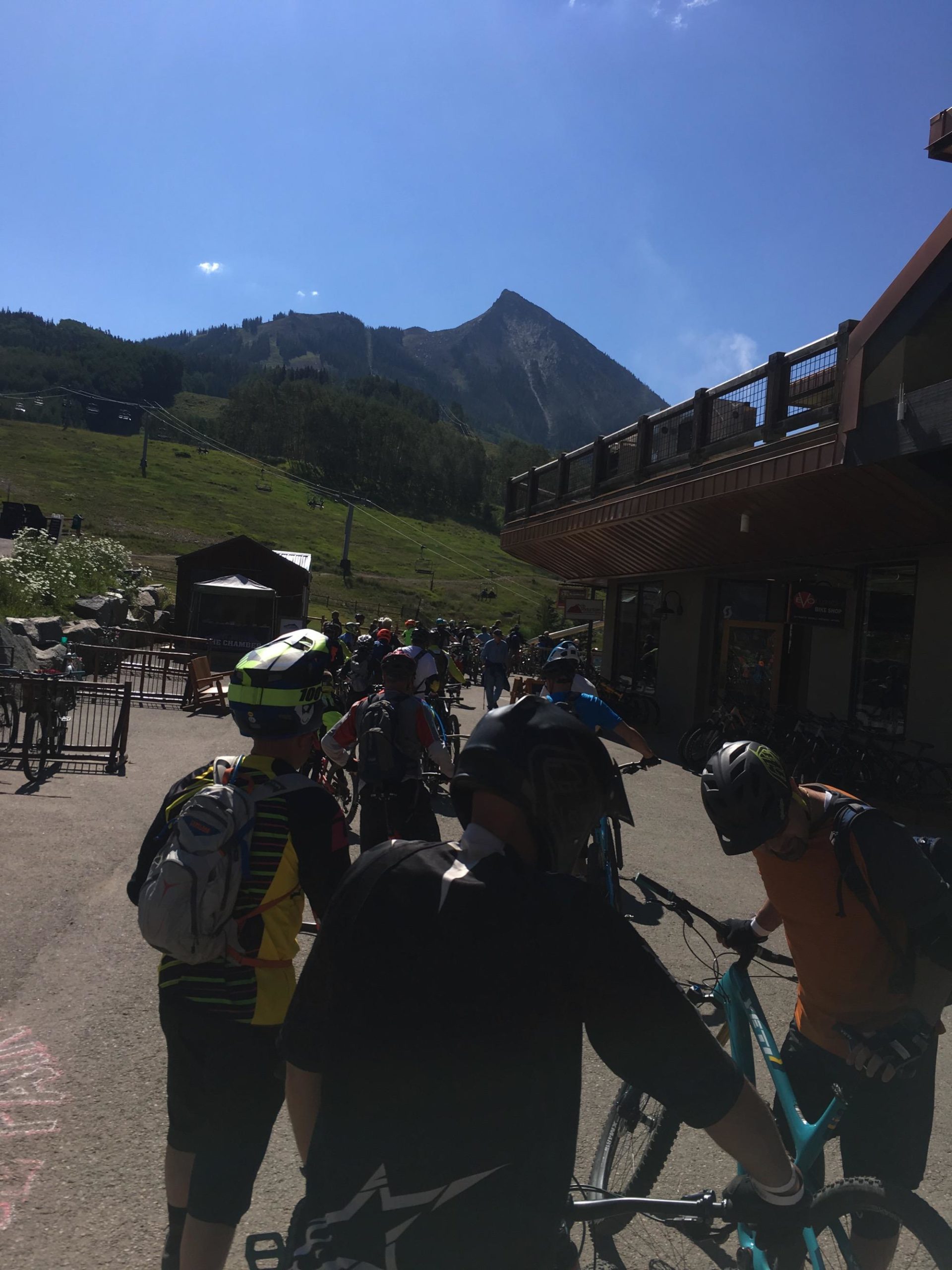 A group of mountain bikers in helmets and gear are gathered near a bike rental area, with a scenic mountain backdrop under a clear blue sky. They are preparing their bikes, some adjusting equipment, while others stand in a line waiting. The area includes buildings that support outdoor activities, indicating a vibrant cycling community. Evolution Bike Park at Crested Butte Mountain Resort mountain bike trail.