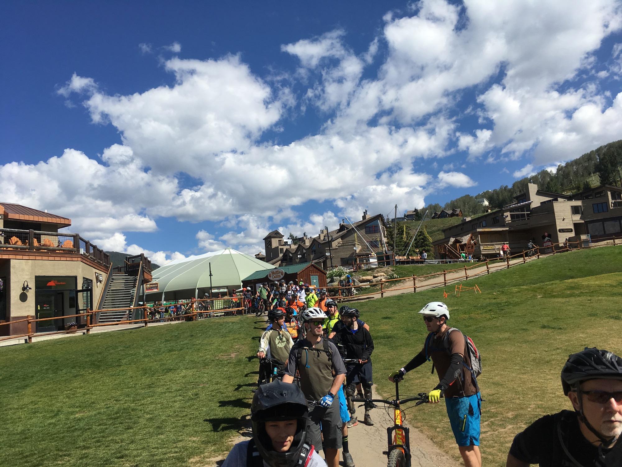 A crowded line of mountain bikers waiting for access to a bike trail in a recreational area, with mountains and blue skies in the background. Various buildings and amenities are visible, along with grass and a dirt path in the foreground. Evolution Bike Park at Crested Butte Mountain Resort mountain bike trail.
