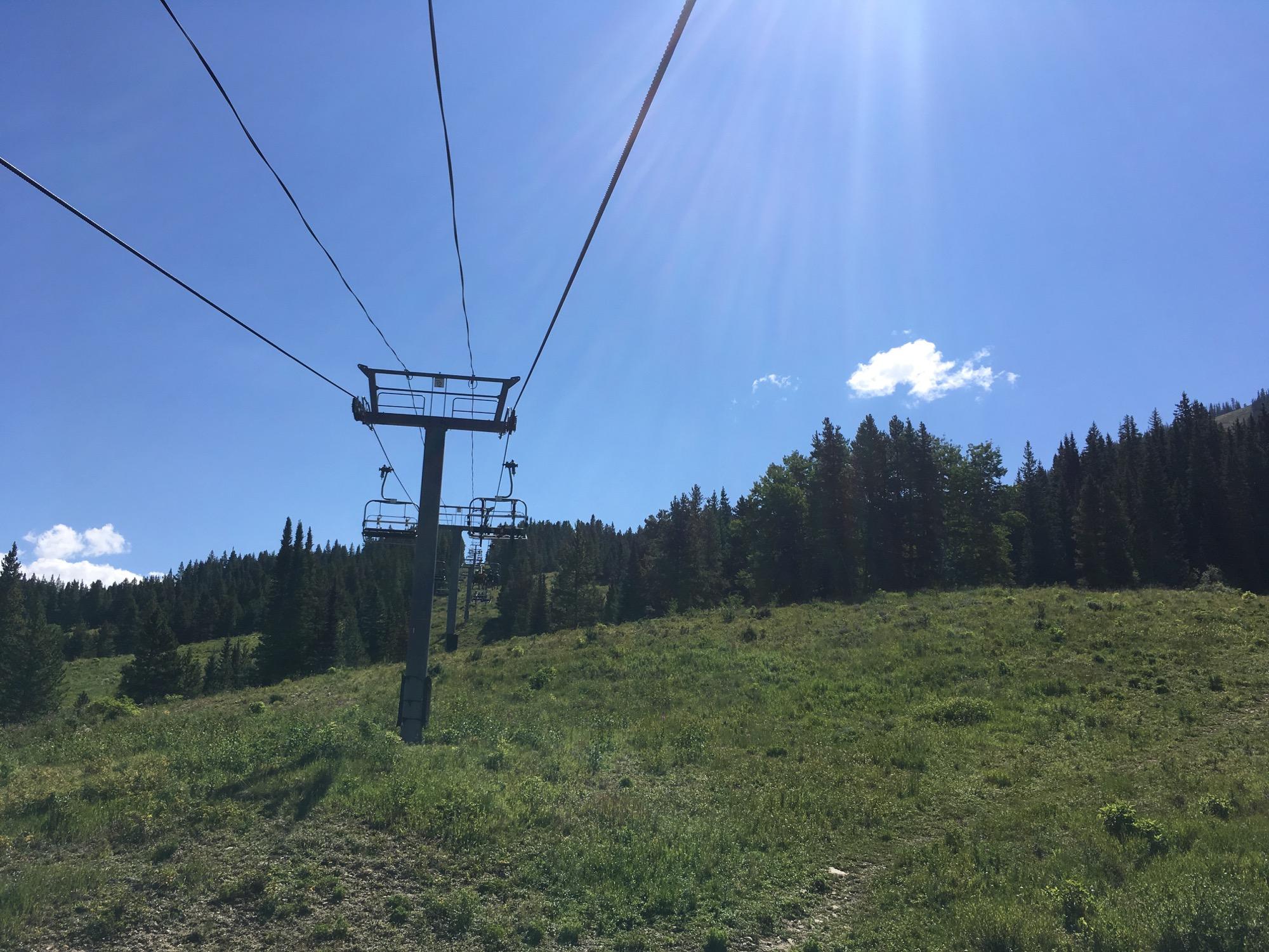 A chairlift extends towards the blue sky, with green grassy hills and pine trees in the background. The sunlight casts a bright glow over the landscape, creating a serene outdoor setting. Evolution Bike Park at Crested Butte Mountain Resort mountain bike trail.