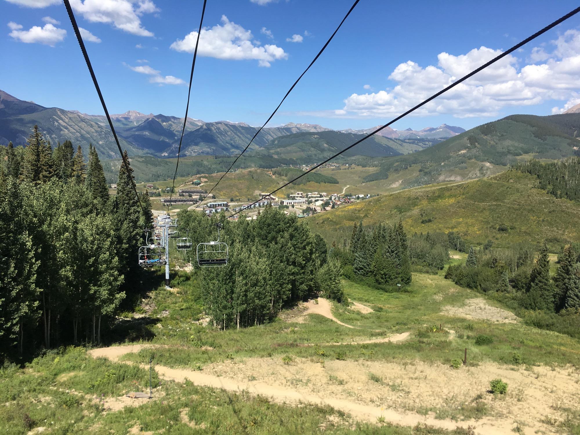 A scenic view from a ski lift, showing green valleys and distant mountain ranges under a blue sky with cloud formations. The lift cable stretches across the foreground, leading toward a town below, surrounded by trees and rolling hills. Evolution Bike Park at Crested Butte Mountain Resort mountain bike trail.