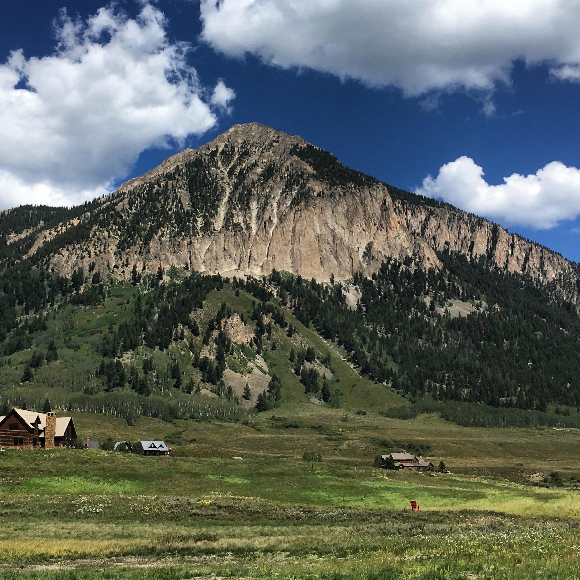 A scenic view of a rugged mountain peak rising against a bright blue sky with fluffy white clouds. In the foreground, a grassy meadow is dotted with wildflowers, and two rustic wooden cabins can be seen at the base of the mountain. The landscape features lush greenery, showcasing the natural beauty of the area. Tony