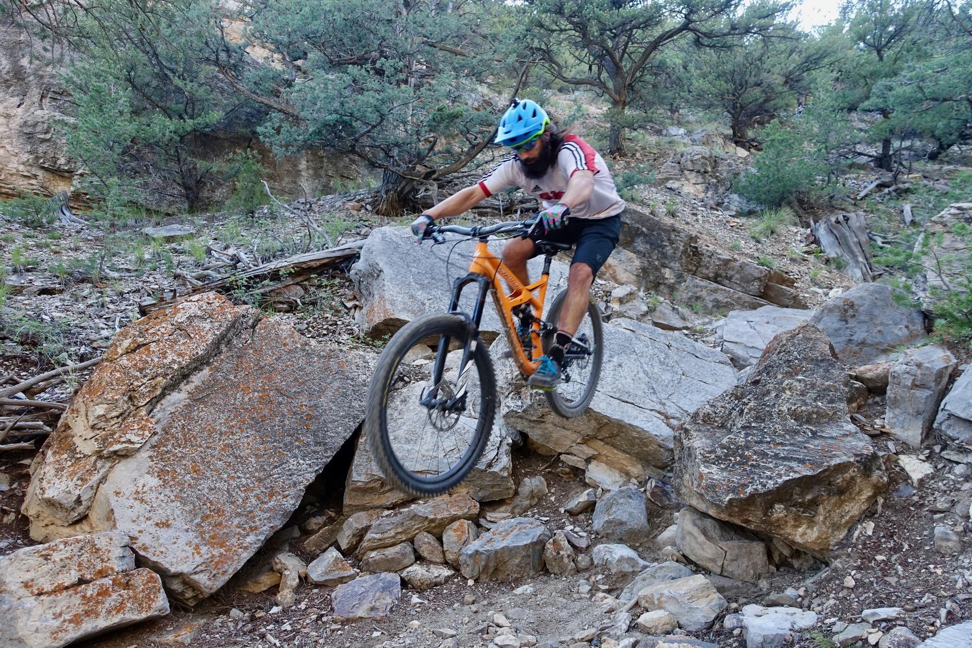 A mountain biker in a blue helmet and light shirt jumps over a rocky terrain surrounded by trees and shrubs. The cyclist rides an orange bike, showcasing skill and agility as they navigate the challenging rocky path. Cottonwood mountain bike trail.