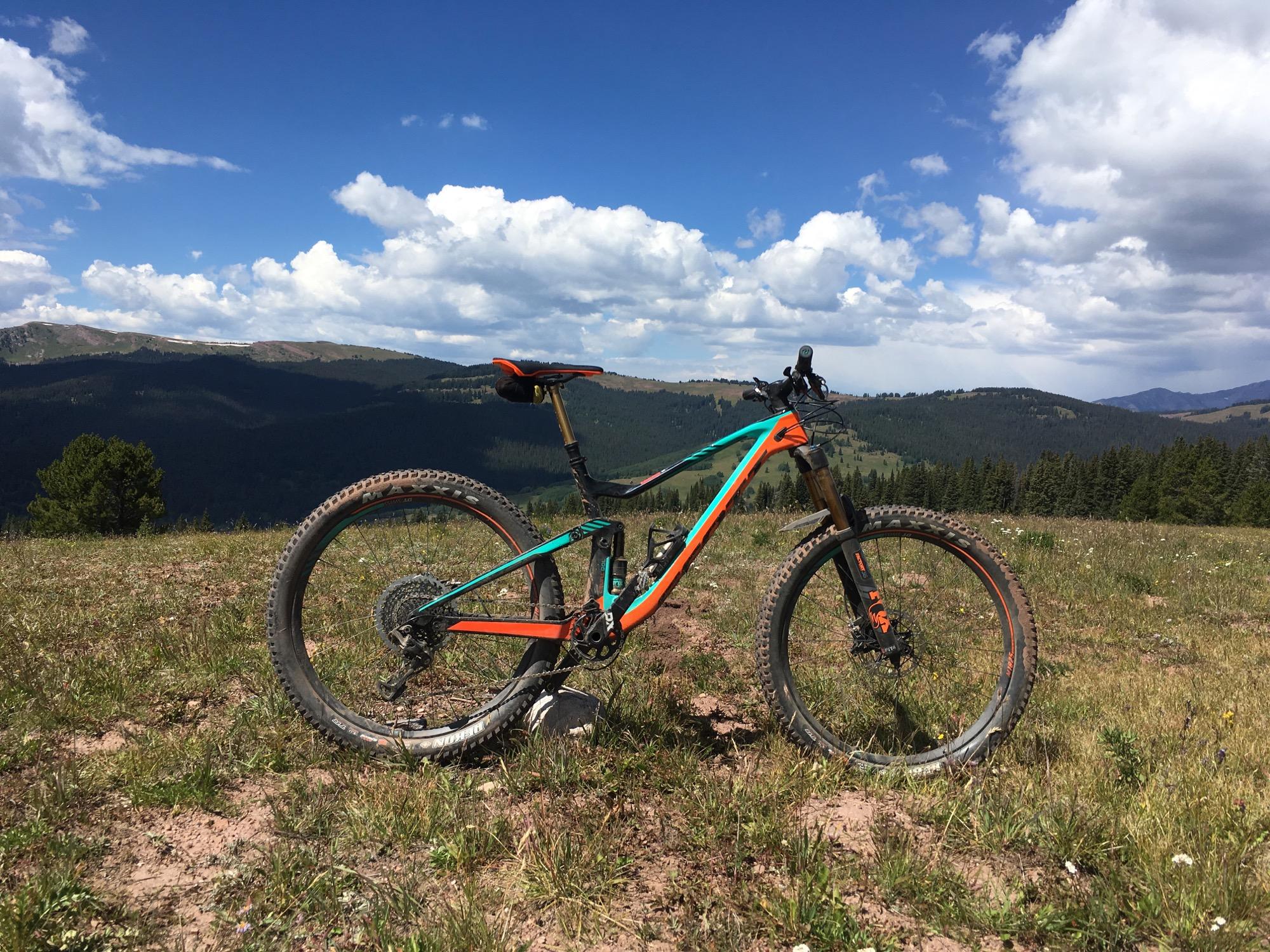 A mountain bike rests on a grassy hilltop, set against a backdrop of rolling hills and a blue sky with scattered clouds. The bike features a vibrant design with orange and teal colors, and its tires are muddy, suggesting it has been used on rugged trails. The scene captures the beauty of nature and the spirit of outdoor adventure. Reno / Flag / Bear / Deadman Loop mountain bike trail.