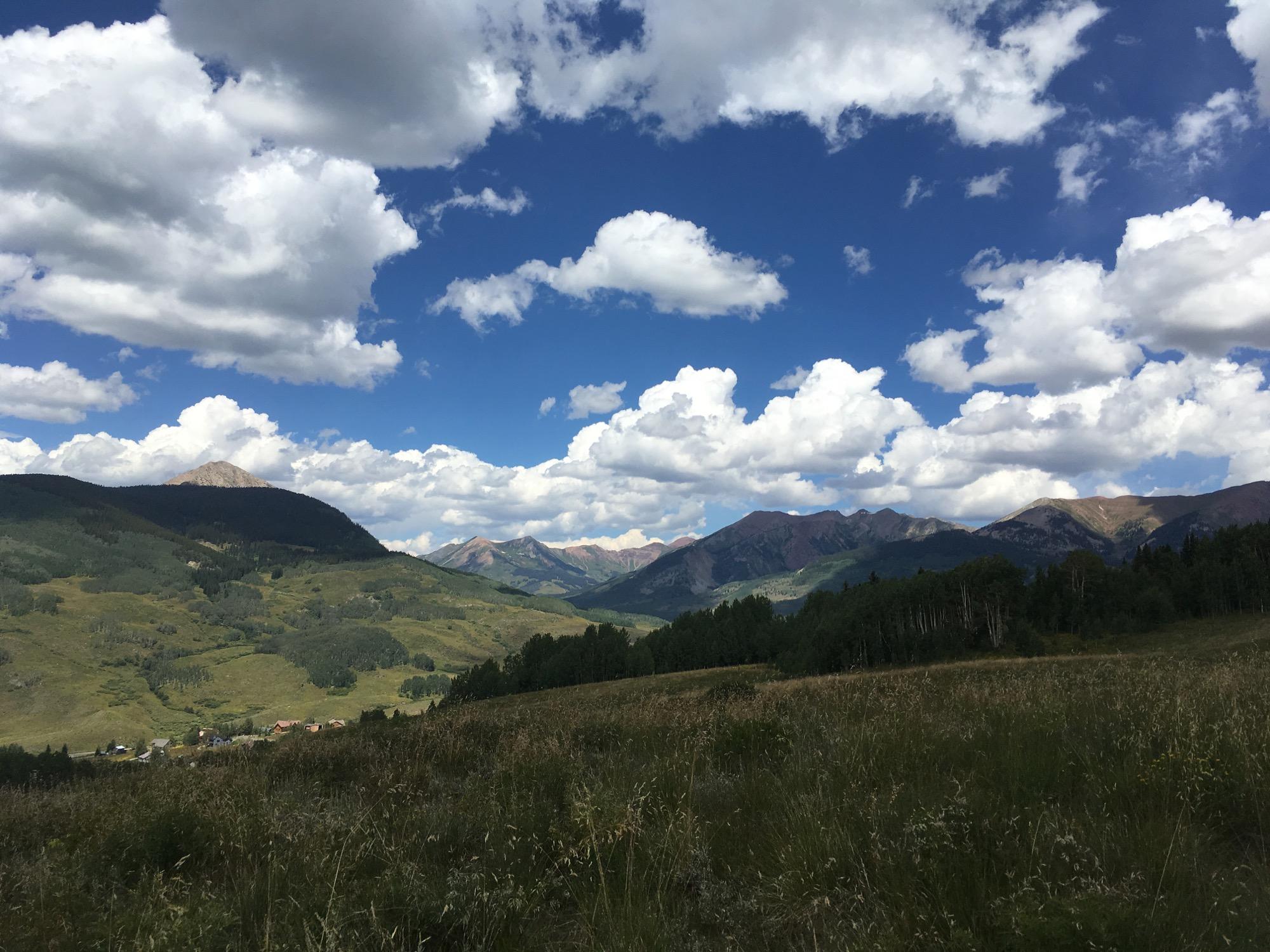 A scenic mountain landscape featuring rolling hills, lush green vegetation, and a partly cloudy blue sky with large, fluffy clouds. In the background, rugged mountains rise against the horizon, showcasing various shades of green and brown. A small town is visible in the valley below. Columbine mountain bike trail.