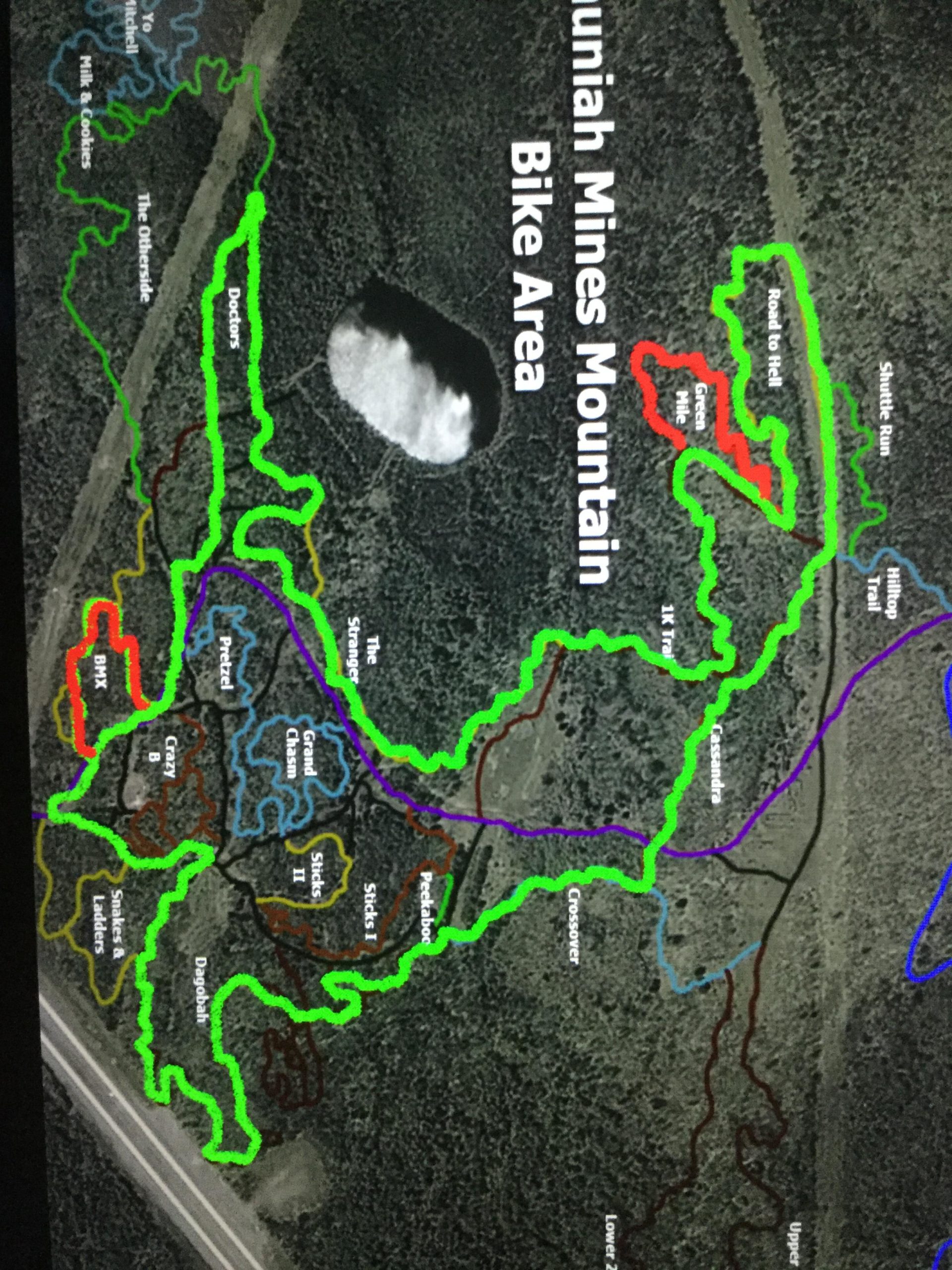 Map of the Huniad Mines Mountain Bike Area, showing various trails and paths in different colors, with trail names labeled. The area includes green, blue, red, and purple trails, along with an aerial view of the surrounding landscape. Shuniah Mines mountain bike trail.
