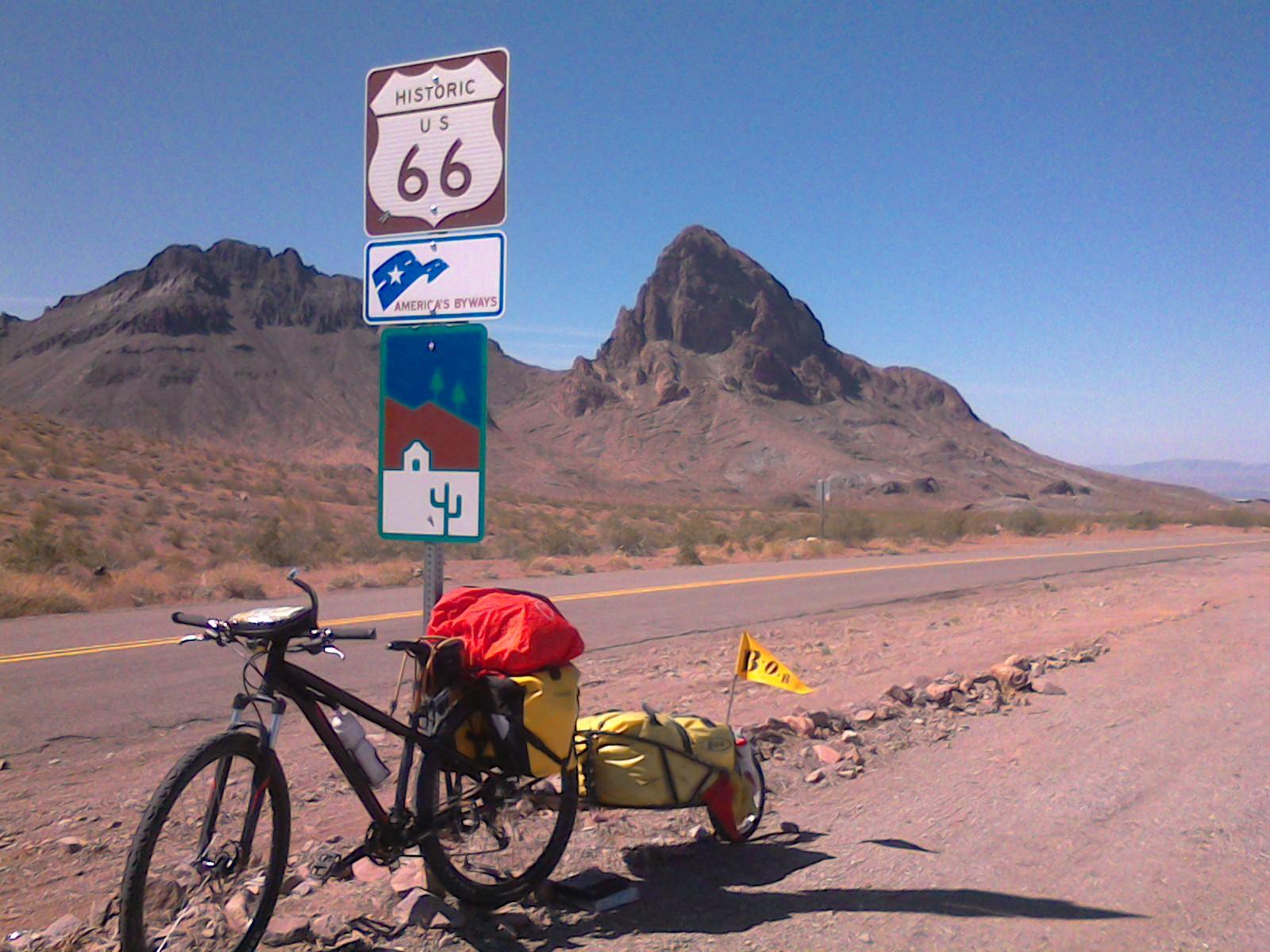 Specialized Hardrock Sport Disc 29er: A bicycle with a yellow and red load is parked beside a Historic U.S. Route 66 sign in a desert landscape, featuring rocky hills in the background and clear blue sky.